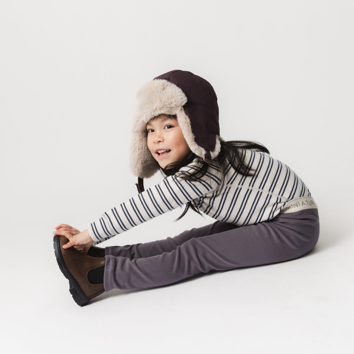 Barn iført MATLEEMA letvægts-fleece-tights i gråblå med synlig MINI A TURE-logolinning, stylet med en stribet langærmet trøje, vinterstøvler og en fluffy pilothat, siddende og strakt fremad mod hvid baggrund.