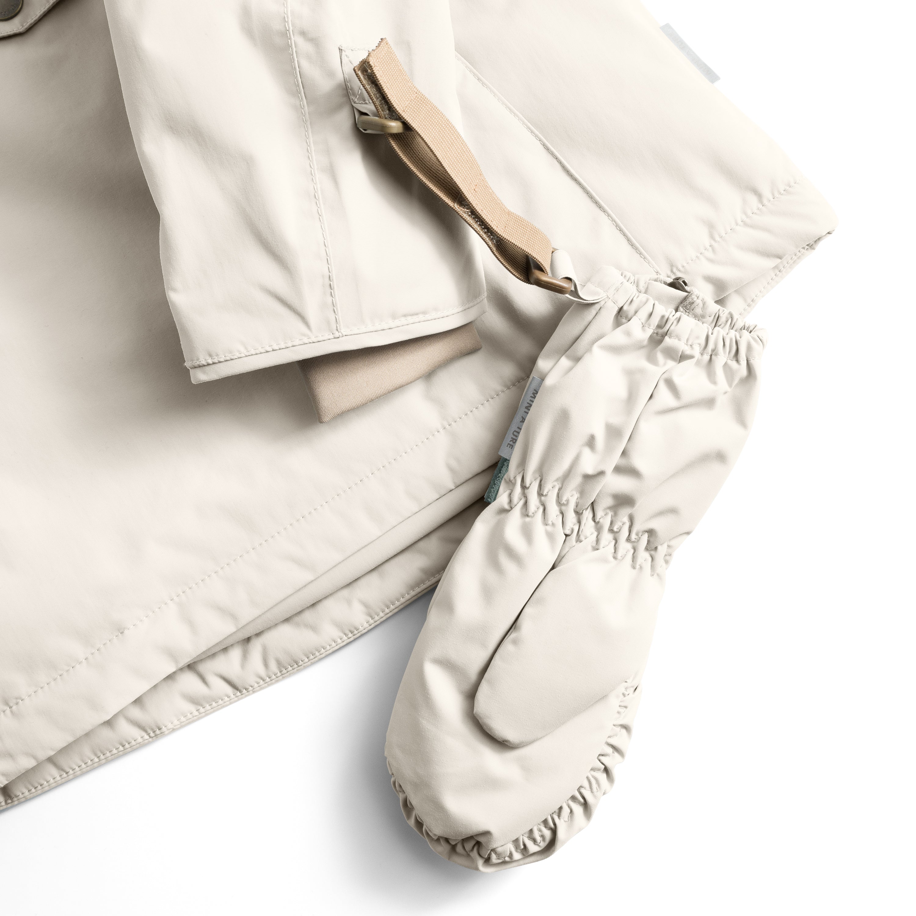 Et par matchende fleeceforede luffer i lys beige er placeret foran en jakke i samme farve, set fra oven. Den sandbeige velcrostrop med messingspænde på jakkens ærme viser, hvordan lufferne kan fastgøres til overtøjet. Den rynkede elastik ved luffernes håndled og fingerspidser samt MINI A TURE-mærket på siden er tydeligt synlige.