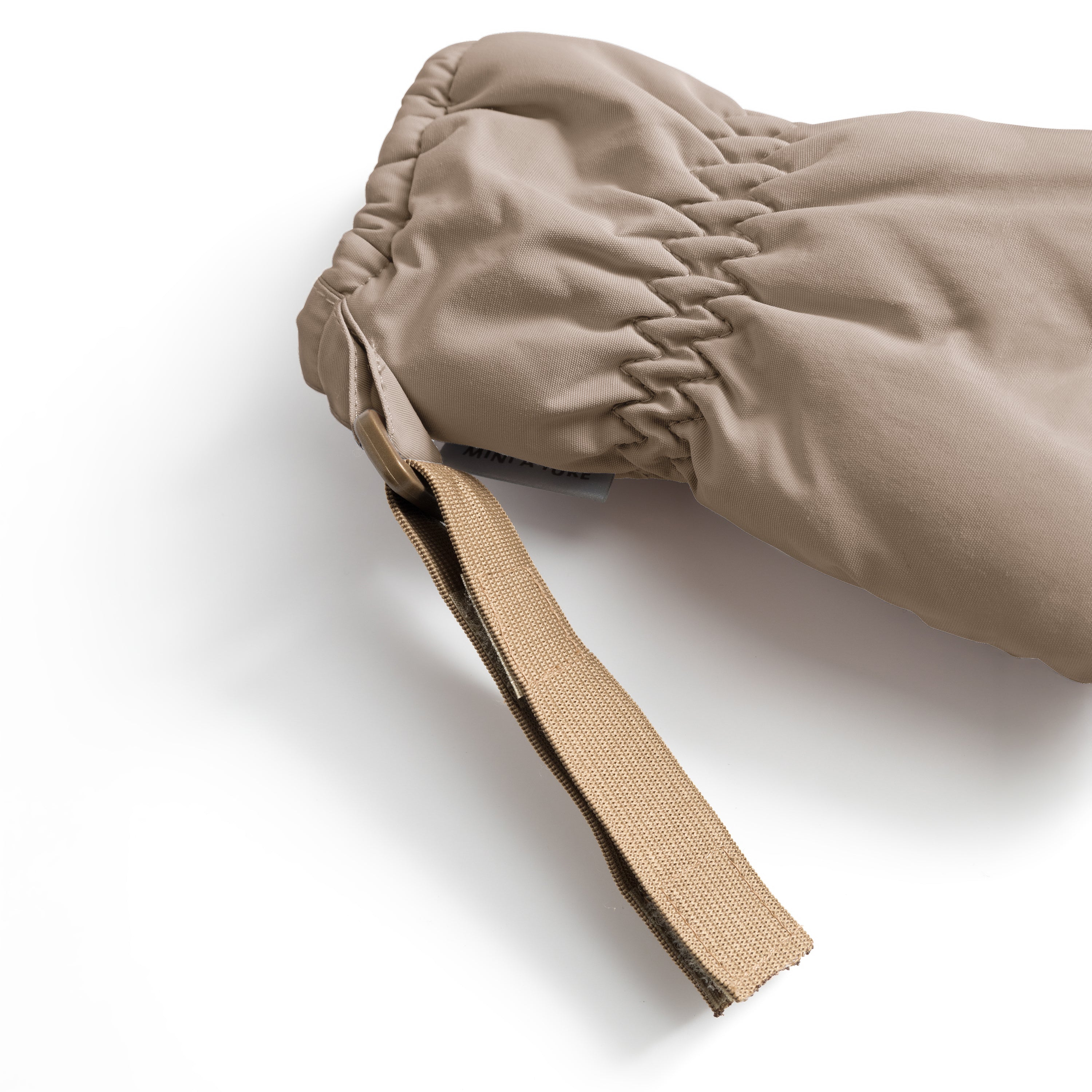 Nærbillede af håndledsdelen på en MATCORDT-fleeceforet luffer i varm sandbeige. Billedet er taget skråt ovenfra mod en hvid baggrund, hvor den tætsiddende, rynkede elastik langs manchetten og den løse velcrostrop med messingfarvet spænde er i fokus.