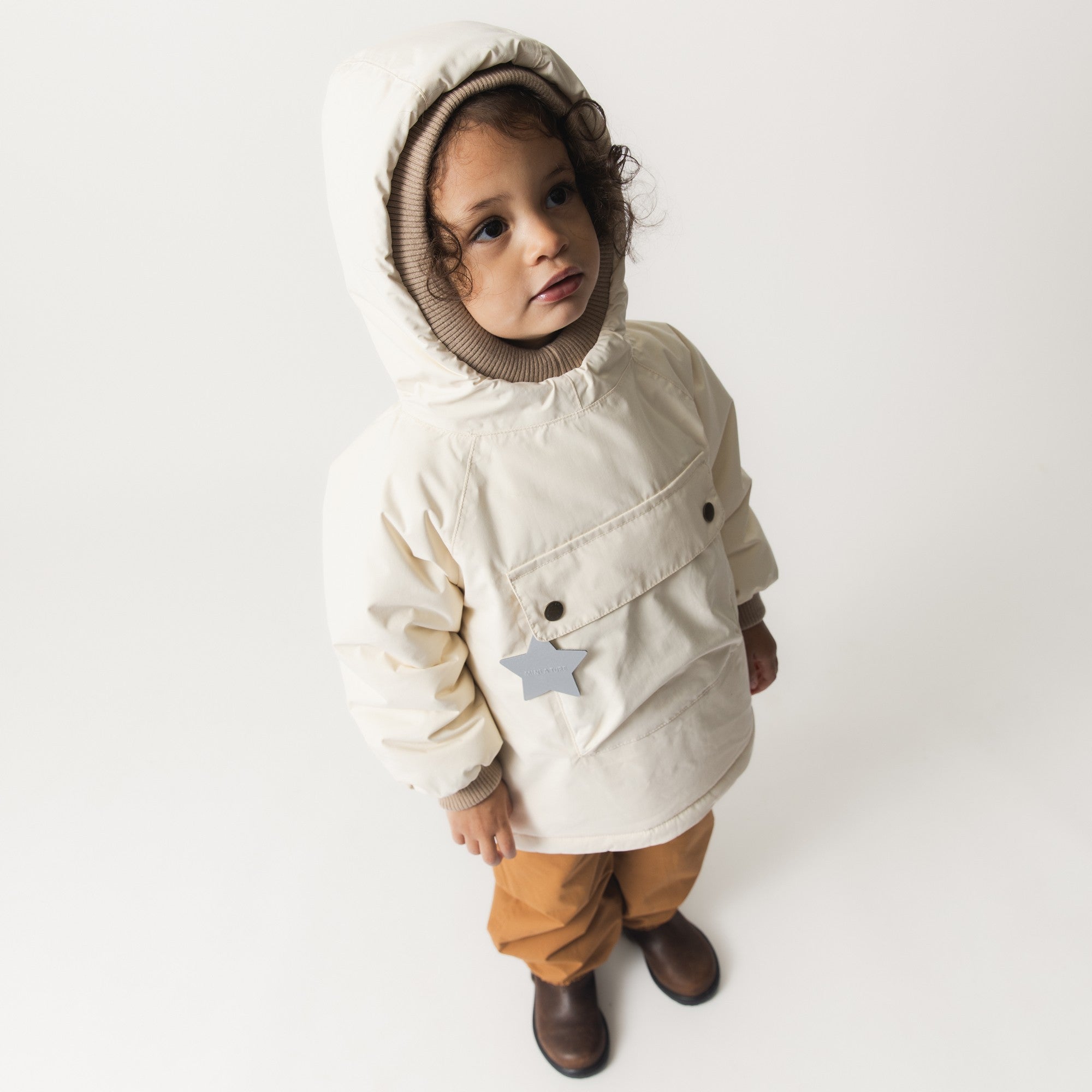 Et lille barn iført en MATBABYWEN fleeceforet vinteranorak i cremefarvet yderstof, fotograferet forfra fra et let fugleperspektiv. Den rummelige hætte med beige ribkant omslutter barnets ansigt, frontlommen er lukket med to mørkegrå trykknapper og har en grå stjernerefleks, og de beige ribmanchetter er synlige ved håndleddene.