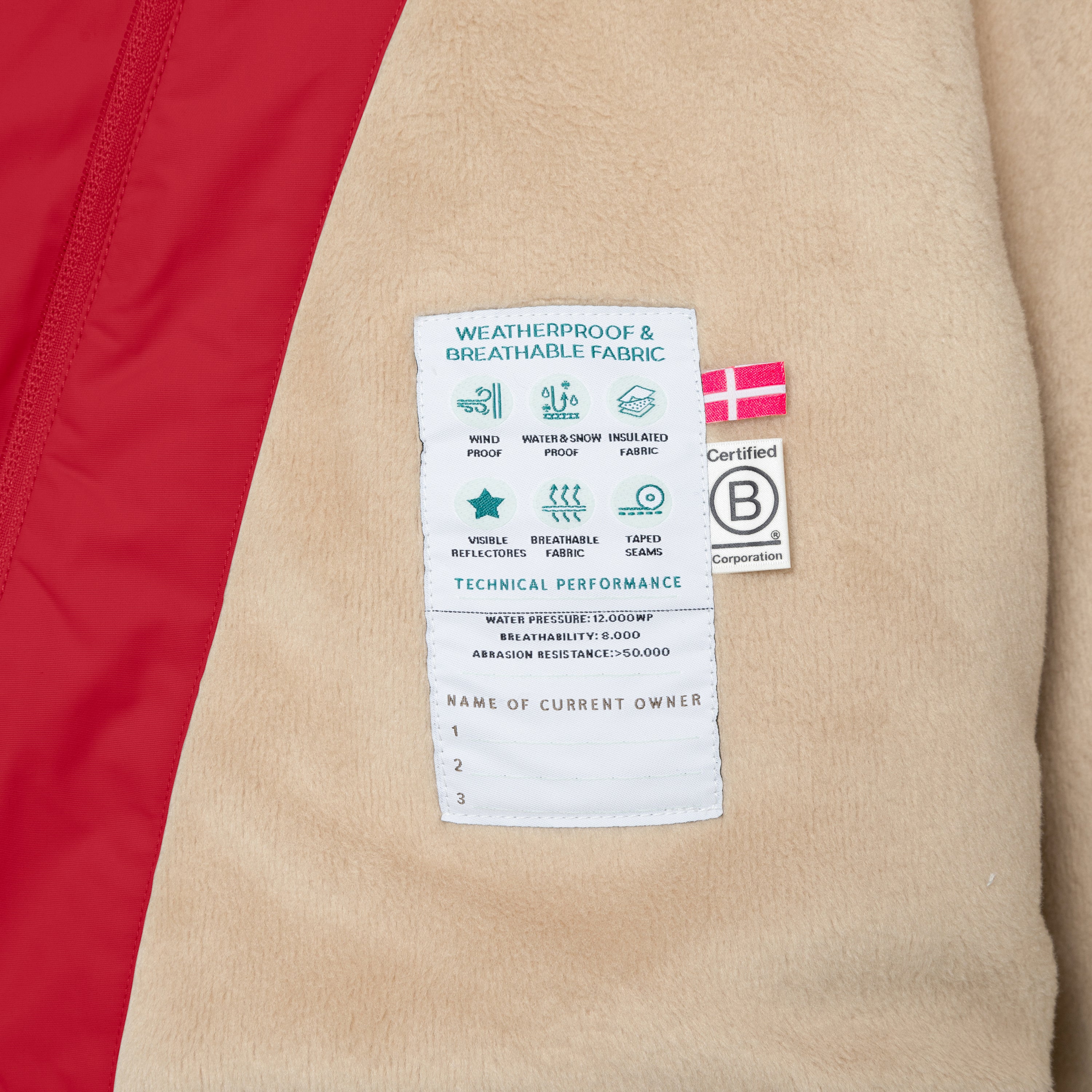 Nærbillede af indersiden af den røde MATWANG-vinterjakke med fleecefor, hvor det beige fleecefór og en indvendig performanceetiket med tekniske specifikationer er synlige. Derudover ses et dansk flag-label og B Corp-certificeringsmærket.