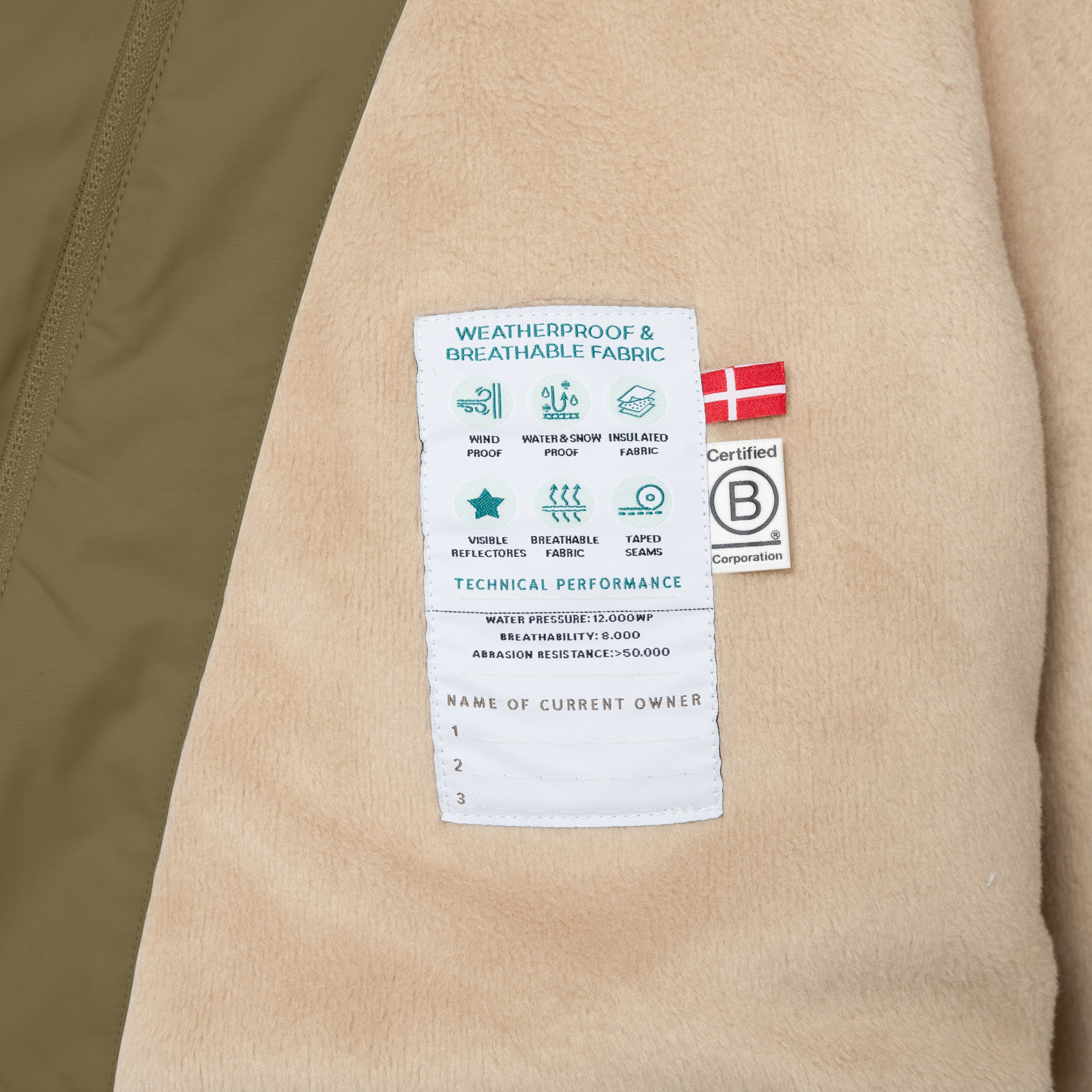 Nærbillede af den indvendige performance-etiket på en MATWANG fleeceforet vinterjakke i olivengrøn. Etiketten er syet fast på det bløde, beige fleecefór og viser tekniske specifikationer som vandtryk på 12.000 WP, åndbarhed på 8.000 og slidstyrke over 50.000. Desuden er der tre navnefelter, så jakkens ejere kan noteres gennem dens levetid.