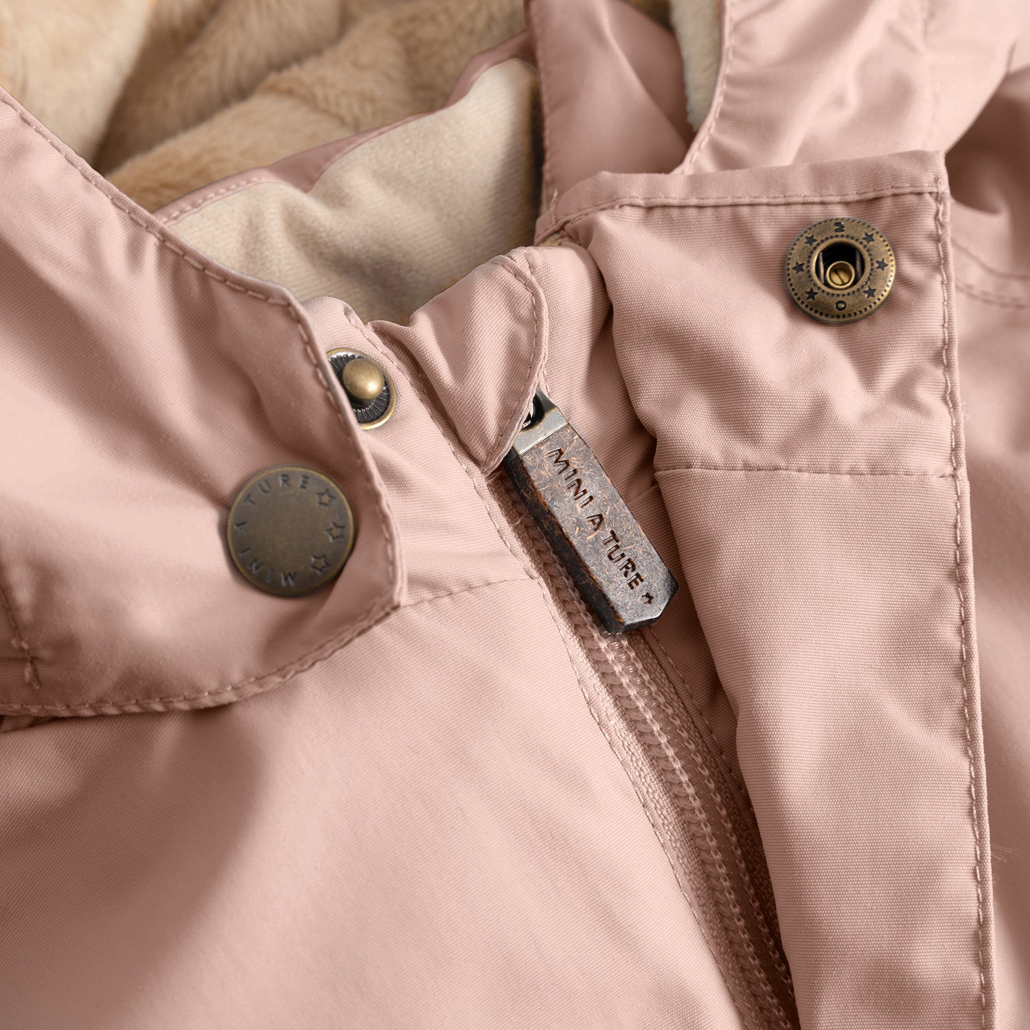 Nærbillede af lynlåsen på MATWALLYs fleeceforede vinterjakke i støvet rosa, hvor MINI A TURE-lynlåspulleren i metal og de to bronzefarvede trykknapper til hætten er i fokus. Det bløde, beige fleecefor er synligt langs hættens åbning øverst på billedet.