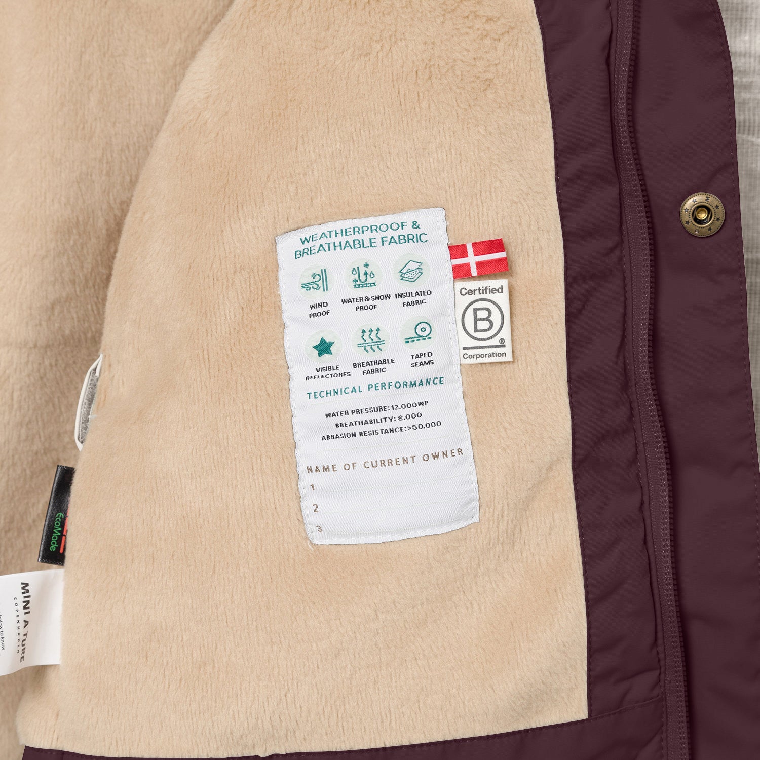 Nærbillede af indersiden af en MATWALLY fleeceforet vinterjakke i mørk blommefarve, hvor den bløde beige fleeceforing viser en hvid performancelabel med tekniske specifikationer, et dansk flag og B Corp-mærket. Til venstre ses MINI A TURE- og EcoMade-mærkerne, og langs kanten er det dybe lilla yderstof med en bronzefarvet trykknap synligt.