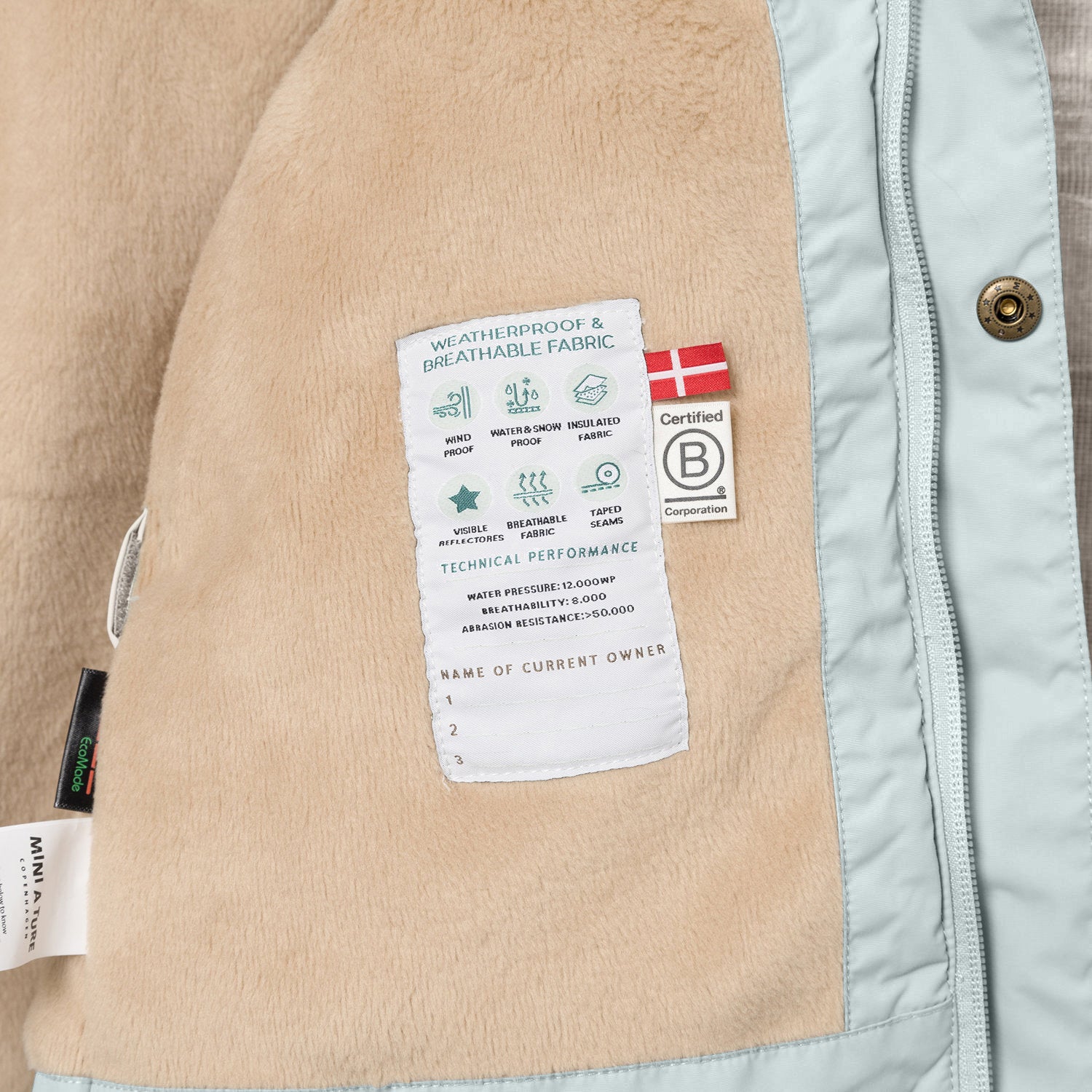 Nærbillede af indersiden af en MATWALLY fleeceforet vinterjakke i lys isblå. Her er den beige fleeceforing åbnet og afslører en hvid performancelabel med tekniske specifikationer samt B Corp-mærket med det danske flag. Til venstre ses MINI A TURE-mærket og en EcoMade-label, og langs kanten er den lyseblå lynlås med en bronzefarvet trykknap synlig.