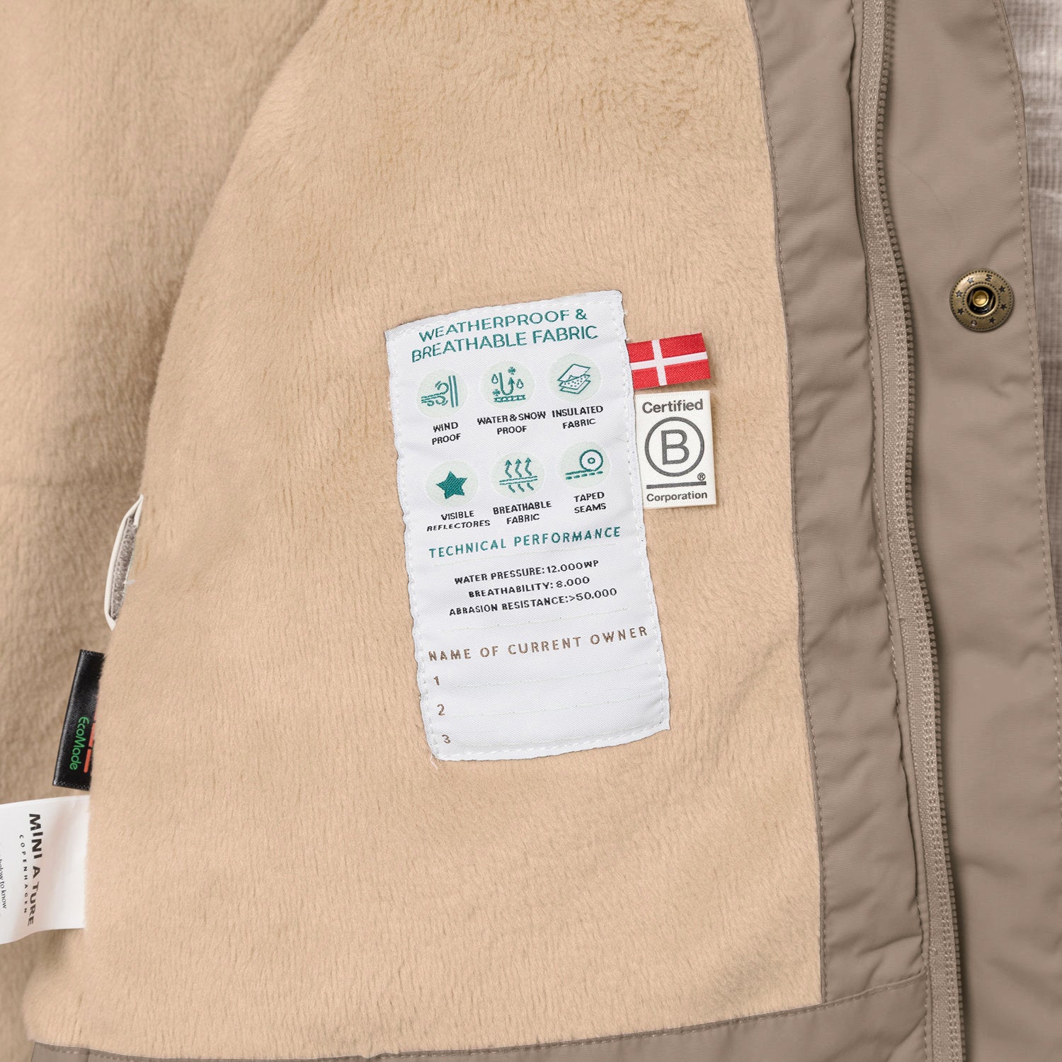 Nærbillede af indersiden af en MATWALLY fleeceforet vinterjakke i sandfarvet brun. Den bløde, beige fleeceforing er åbnet, så man kan se en hvid performancelabel med tekniske specifikationer samt B Corp-mærket med det danske flag. Til venstre ses MINI A TURE-mærket og en EcoMade-label, der er syet fast i sømmen.
