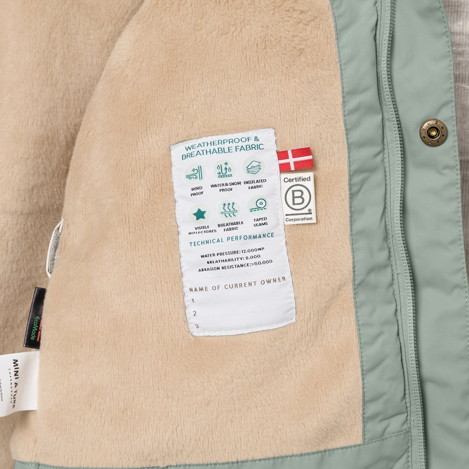 Nærbillede af indersiden af en MATWALLY fleeceforet vinterjakke i støvet mintgrøn, der viser den bløde, beige fleeceforing med en hvid performancelabel. På labelen ses tekniske specifikationer, det danske flag og B Corp-mærket. Til venstre er MINI A TURE-mærket og EcoMade-labelen synlige, mens den mintgrønne lynlås og en bronzefarvet trykknap ses til højre.
