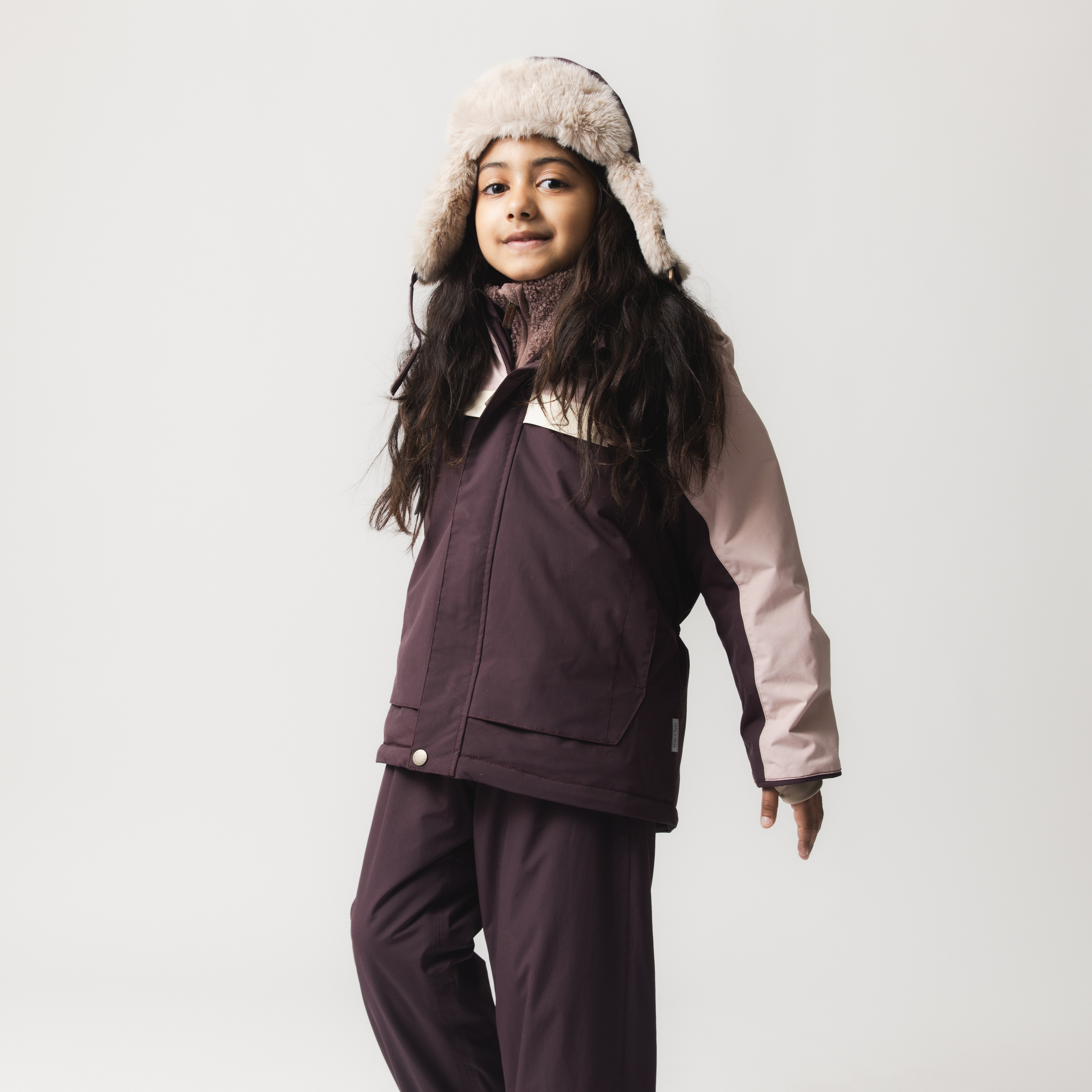 Et barn iført en MATWALDO fleeceforet vinterjakke i et tofarvet design med mørk aubergine på kroppen og lyserøde ærmer, kombineret med matchende mørke vinterbukser og en beige pelsforet hue med øreklapper. Jakken er lukket og viser den bløde fleecekrave ved halsen, mens barnet smiler og vender sig let til siden.