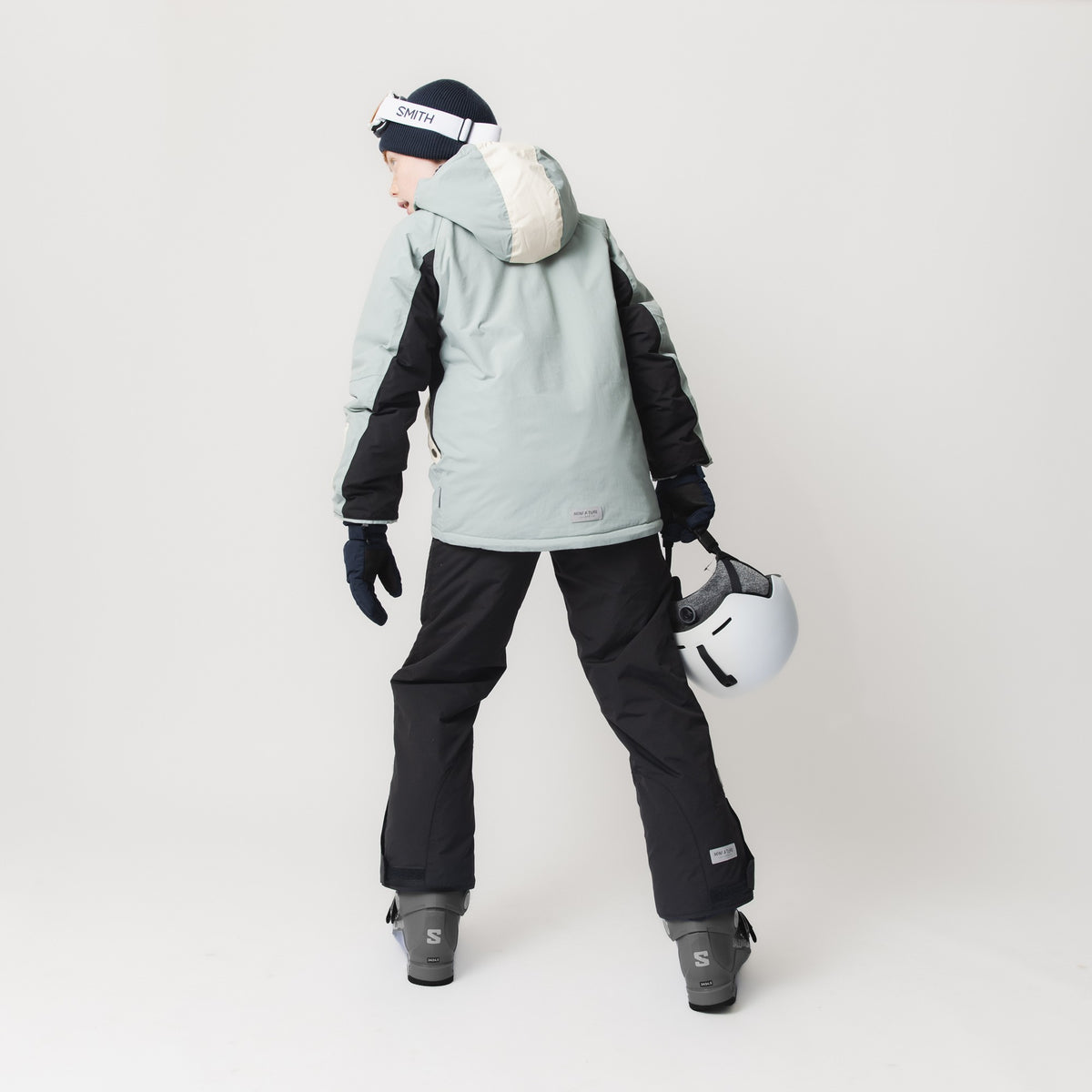 Et barn fotograferet i fuld figur bagfra iført en MATVALONSO skijakke i blågrøn med sorte kontrastpaneler og cremehvidt hætteindlæg, kombineret med sorte skibukser og grå skistøvler. Barnet har mørkeblå skihandsker på, en sort hue med hvide skibriller og holder en hvid skihjelm i hånden.