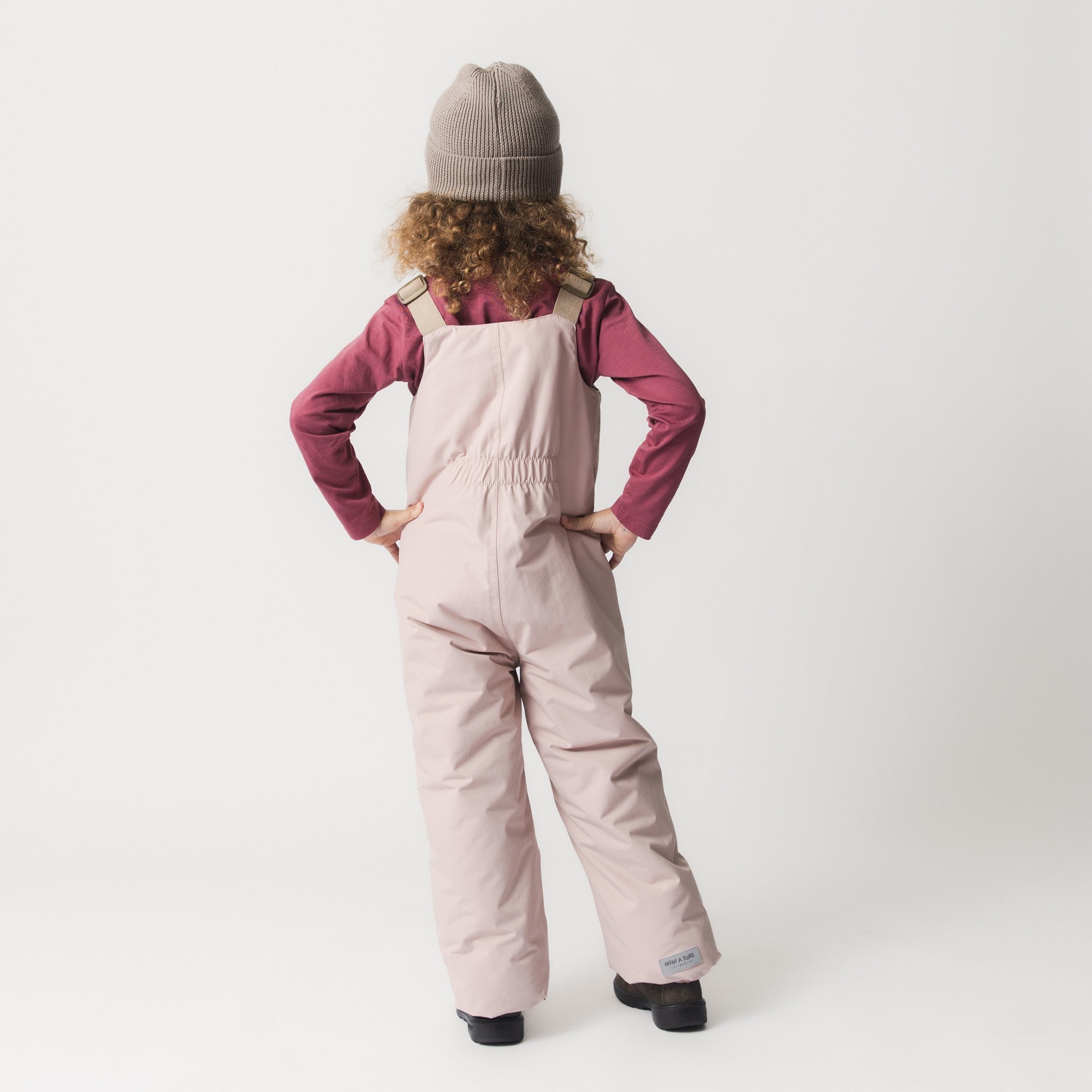 Et barn med krøllet hår og en beige hue set bagfra, iført MATWALENTEX-overtræksbukser i lys pudderrosa med beige seler og elastik i taljen. Bukserne er stylet over en mørkerød langærmet trøje og mørke vinterstøvler.