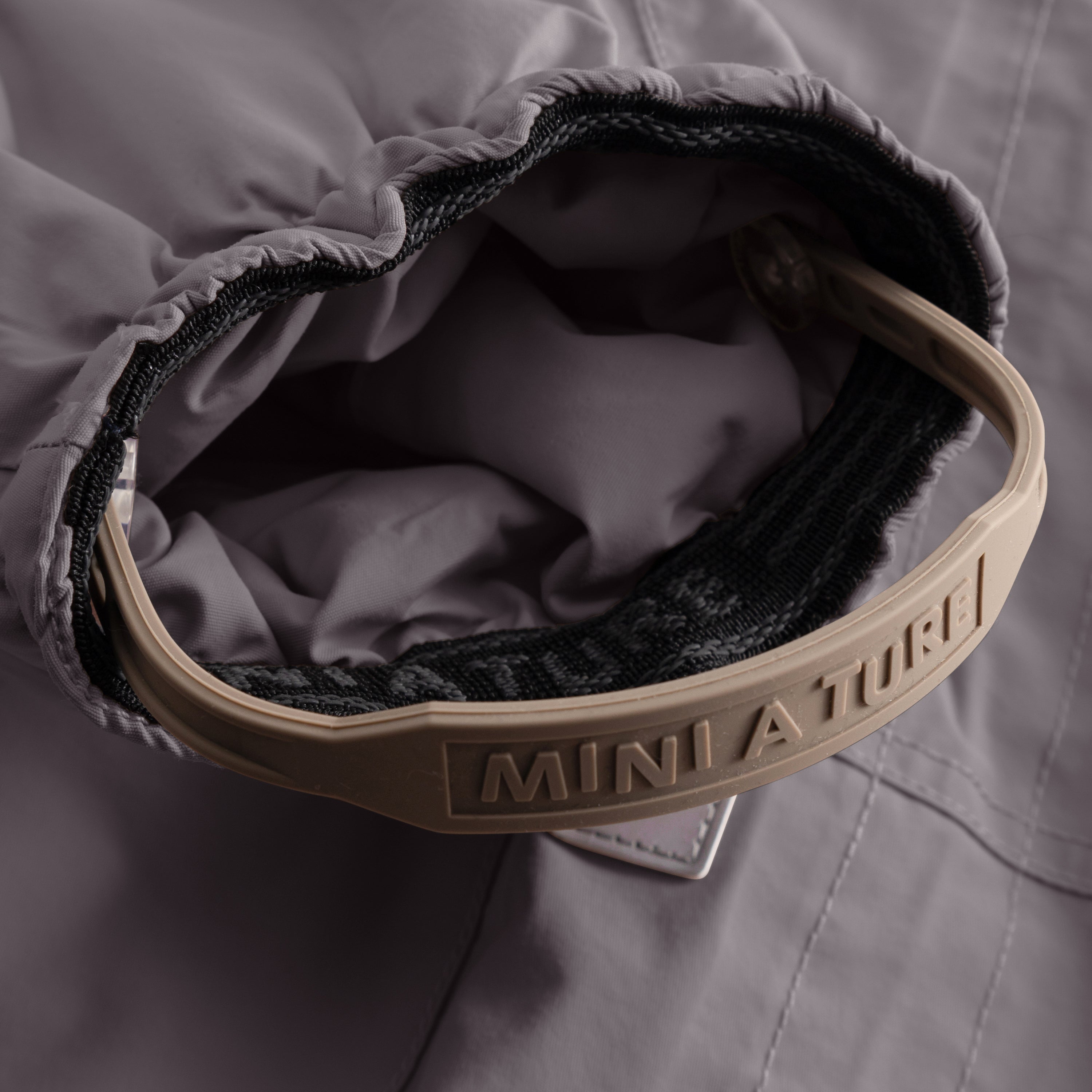 Nærbillede af den åbne forlomme på MATWISTI-fleeceforede flyverdragt i grålilla, hvor det beige MINI A TURE-mærkede lynlåsgreb og det mørke fleecefór inde i lommen er tydeligt synligt.