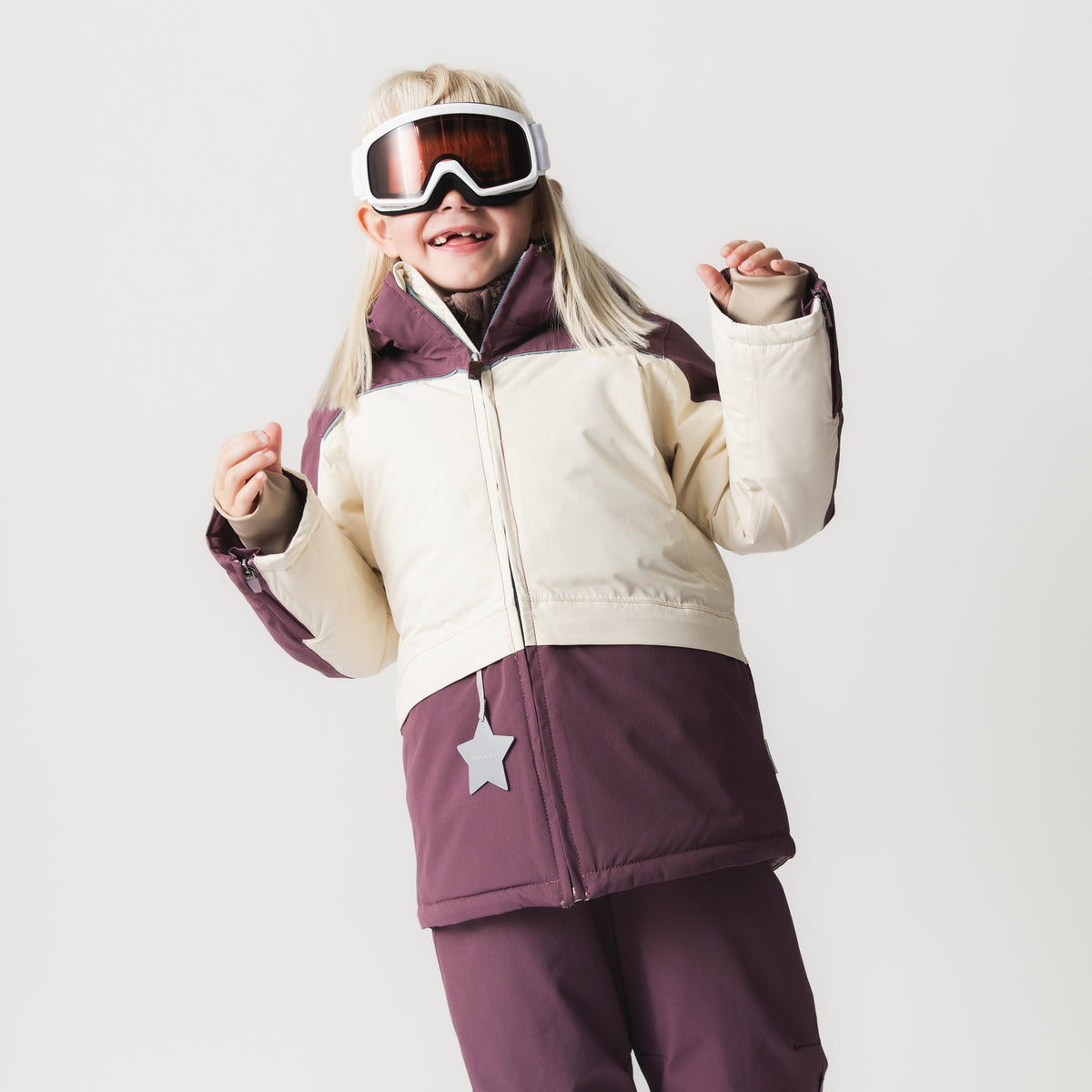 Barn iført MATADANE ski tech-jakke i farveblokdesign med creme og mørkelilla, klar til pisten med skibriller og matchende skibukser.