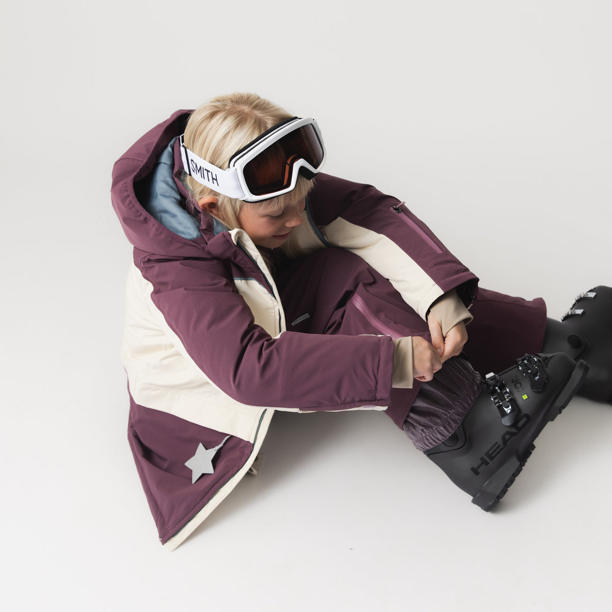 Barnet sidder på gulvet iført MATADANE Ski Tech-jakke i mørkelilla og creme. Med skibrillerne skubbet op på panden spænder det skistøvlerne fast.