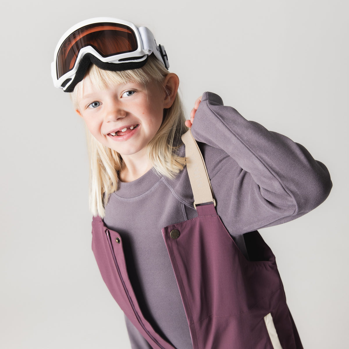 Et smilende barn iført MATADAL skitech-bukser i mørk rosa-lilla med sandfarvede, justerbare seler og lynlås fortil. Bukserne er kombineret med en grå langærmet trøje, og barnet har skibriller på hovedet.