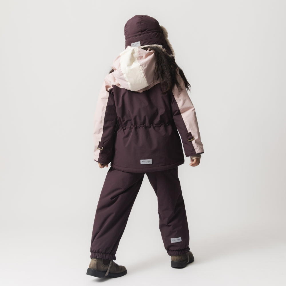 Et barn set bagfra iført en MATWALDO fleeceforet vinterjakke i tofarvet colorblock-design med støvet lyserøde ærmer og cremefarvet hætte kombineret med mørk aubergine på kroppen. Barnet har matchende mørke vinterbukser og en pelsforet vinterhue på. Den indvendige talje er synligt justeret med elastik, og MINI A TURE-mærket ses centreret nederst på ryggen.