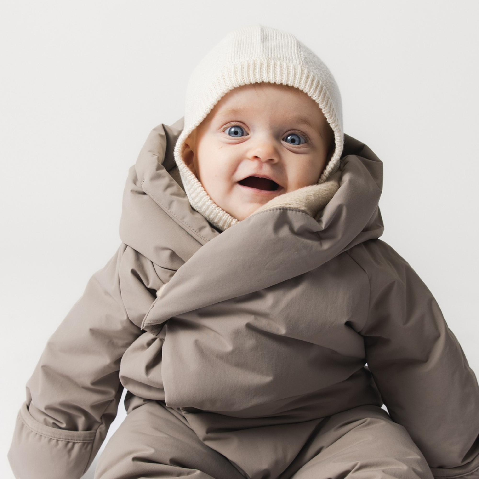 En glad baby iført en MATKEA let elefanthue i offwhite merinould, der dækker ører og nakke tæt ind til huden under en gråbrun vinterdragt.