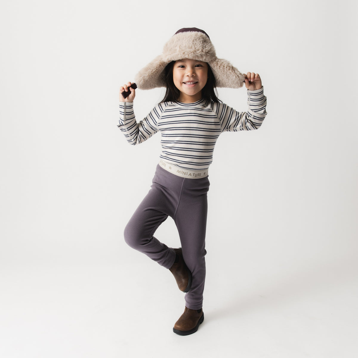 Barn iført MATLEEMA let fleece-tights i støvet lilla med synlig MINI A TURE-logolinning, stylet med en stribet langærmet trøje og en fluffy pilothat, poserende på ét ben mod hvid baggrund.