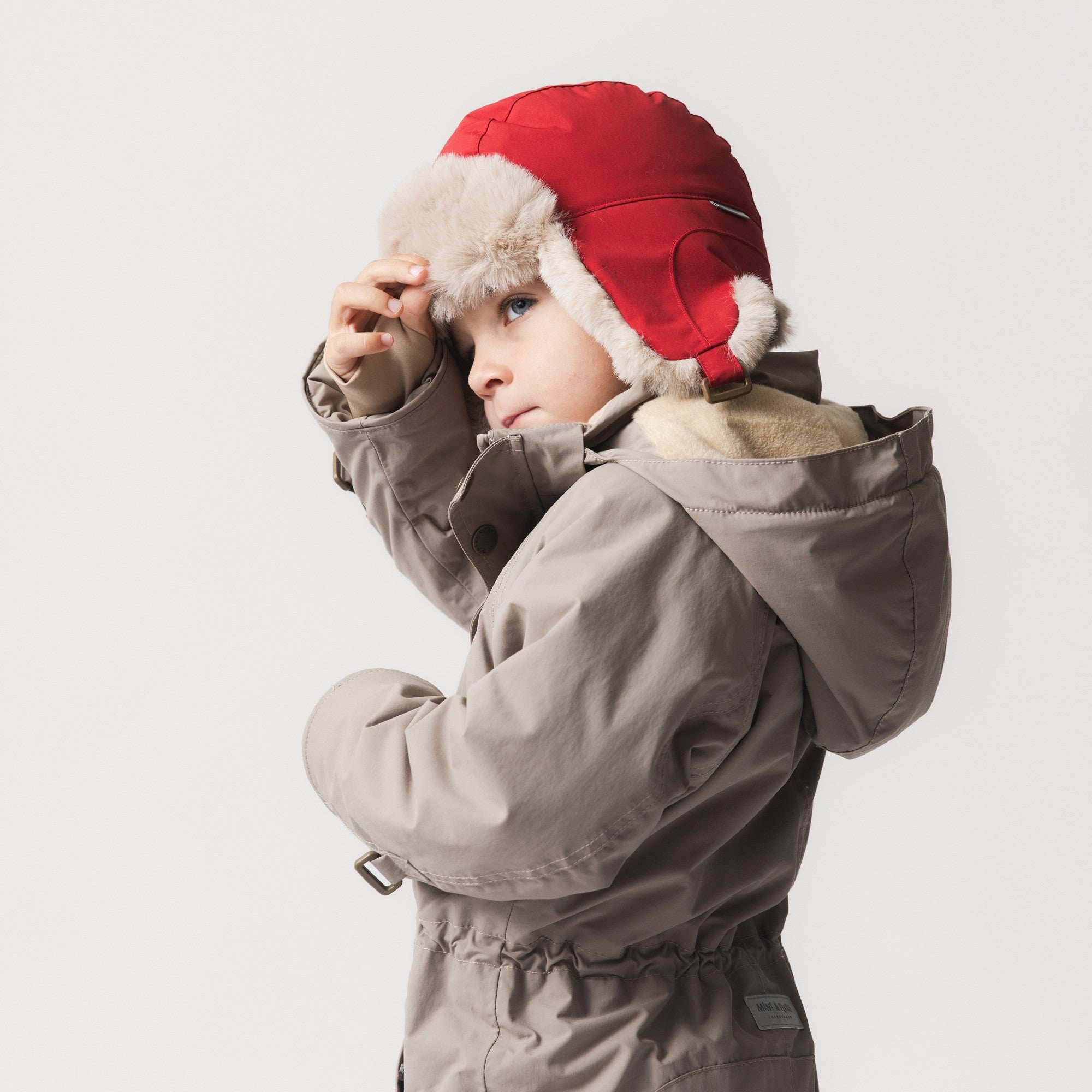 Barn iført MATCRISTER teddyforet vinterhue i knaldrød med cremefarvede øreklapper af teddyfleece. Barnet er fotograferet i halvprofil, mens det retter huen på hovedet, og har en sandfarvet vinterjakke på.