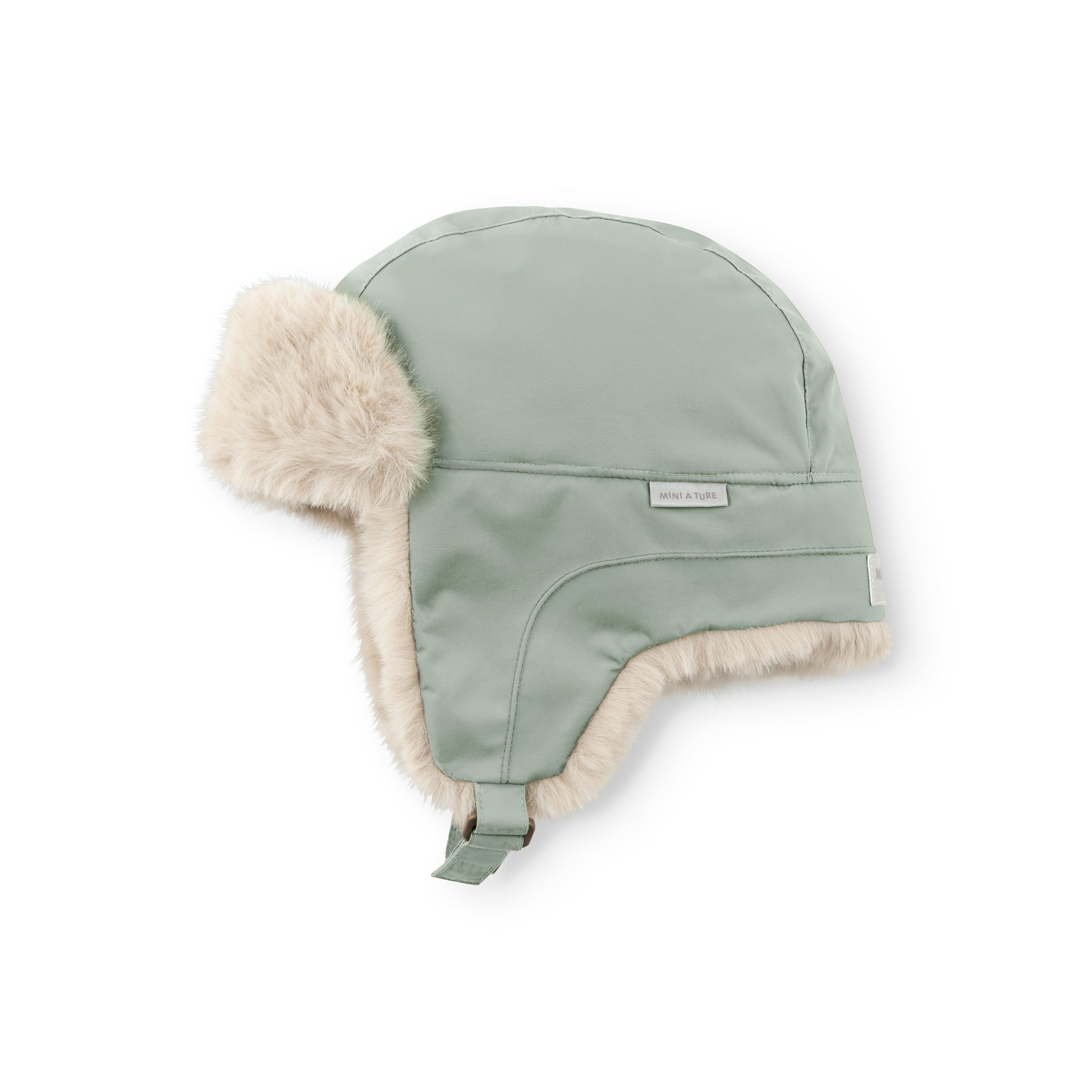MATCRISTER teddyforet vinterhue i støvet mintgrøn, fotograferet fra siden, hvor det cremefarvede teddyfleece ses langs kanten af øreklapperne og øverst på huen. Et MINI A TURE-label sidder på ydersiden, og en matchende grøn velcrostrop hænger ned i siden mod en hvid baggrund.