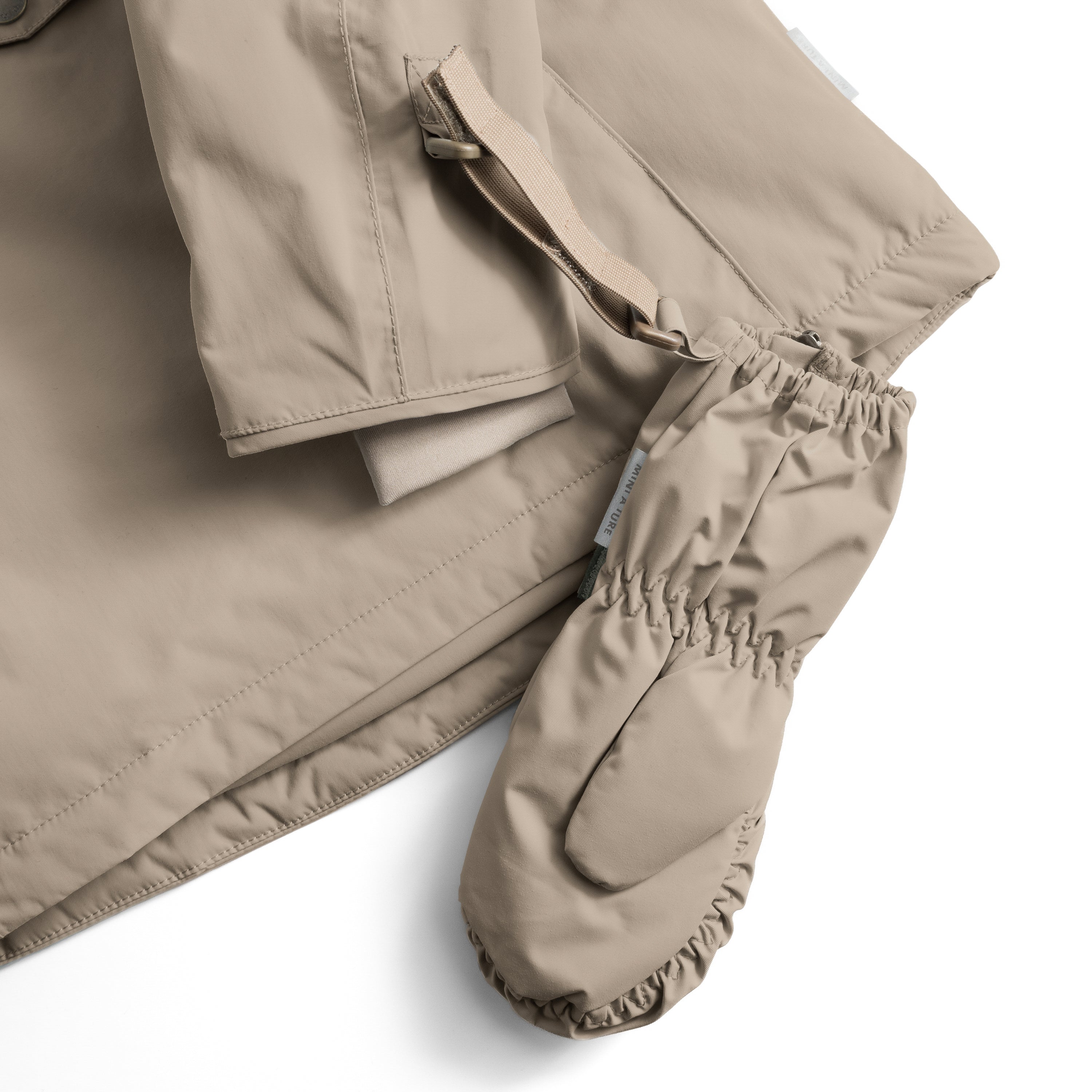 En MATCESAR-fleeceforet luffe i sandfarvet beige ligger op ad en matchende jakke i samme farve. Den brede elastik ved åbningen og MINI A TURE-mærket på luffen er tydeligt synlige, mens velcrostroppen på jakken viser, hvordan luffen kan fastgøres til overtøjet.