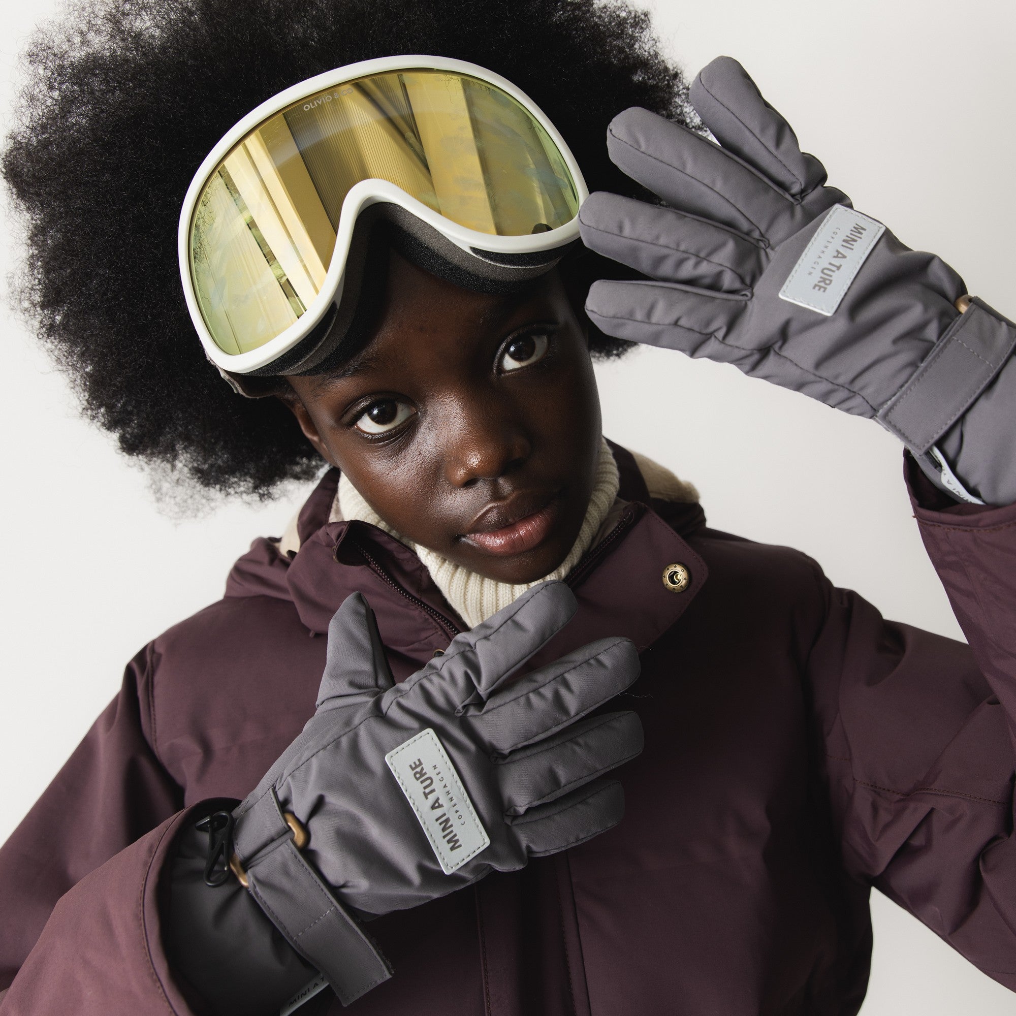 Et barn iført skibriller og en mørkerød vinterjakke har et par grå MATCELIO-handsker på, hvor MINI A TURE Copenhagen-mærket på håndryggen er tydeligt synligt. Det vandtætte yderstof i genanvendt polyamid og den THINSULATE-isolerede konstruktion kommer flot til udtryk i det vinterklare look.