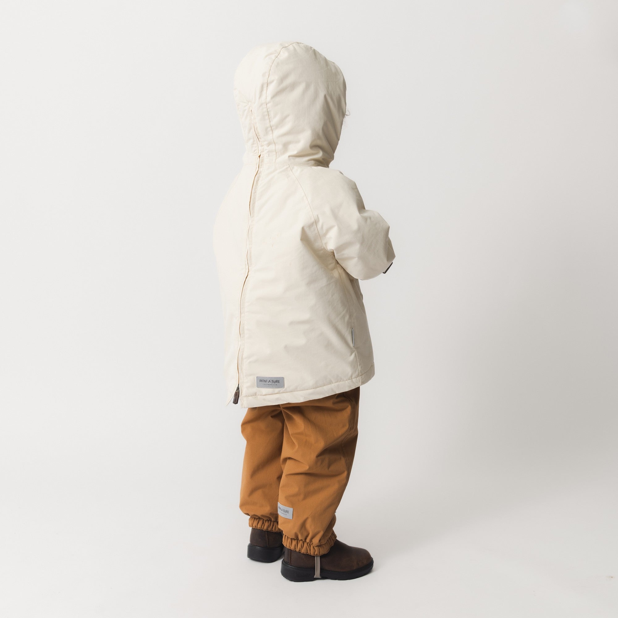 Lille barn set skråt bagfra iført en MATBABYWEN fleeceforet vinteranorak i råhvid med stor hætte og synlig lynlås i ryggen, kombineret med okkerbrune vinterbukser og mørke støvler.