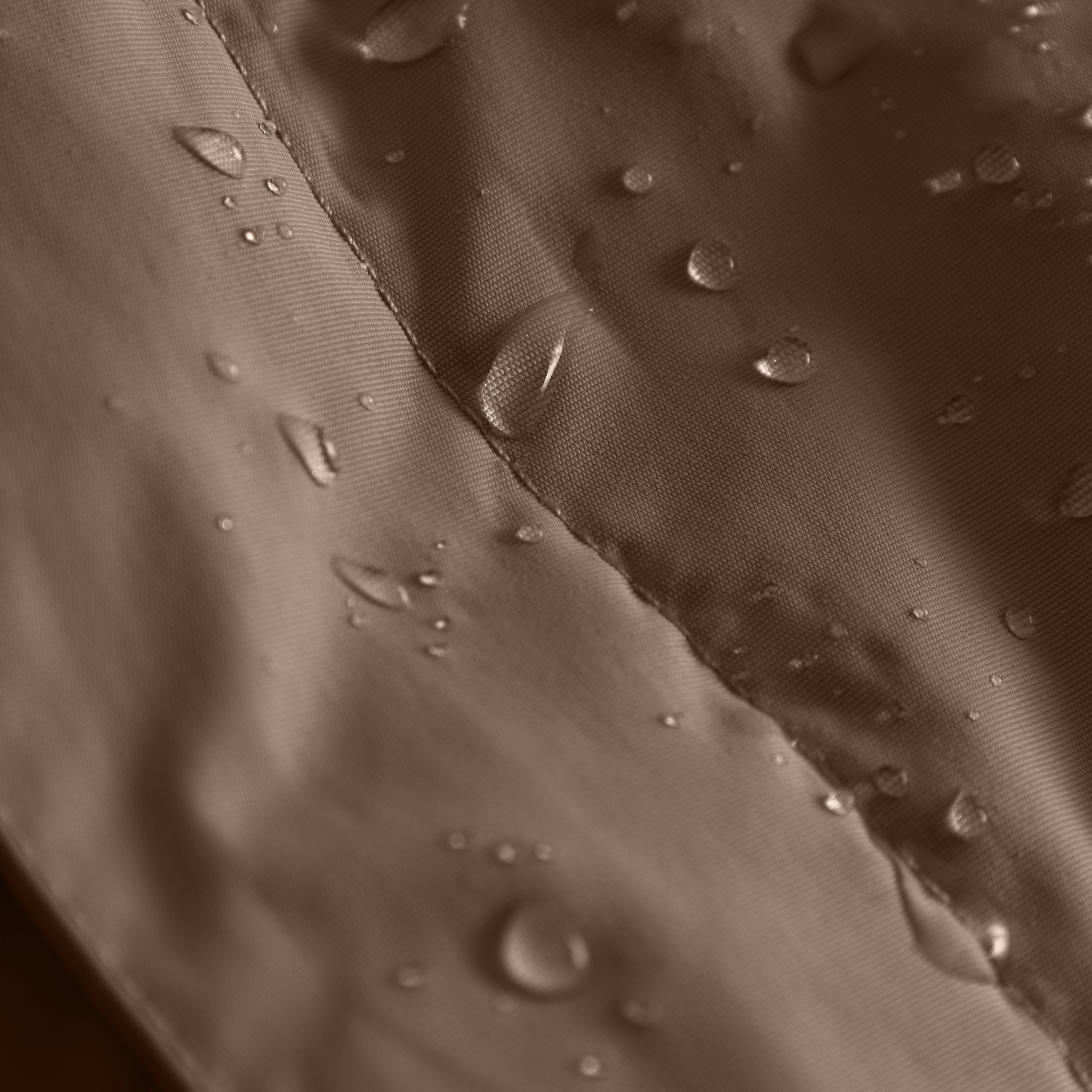 Makrooptagelse af det chokoladebrune yderstof på MATBABYWEN-fleeceforet vinteranorak, hvor vandperler hviler spredt på stoffets overflade på begge sider af en tapetsøm, der løber diagonalt gennem billedet.