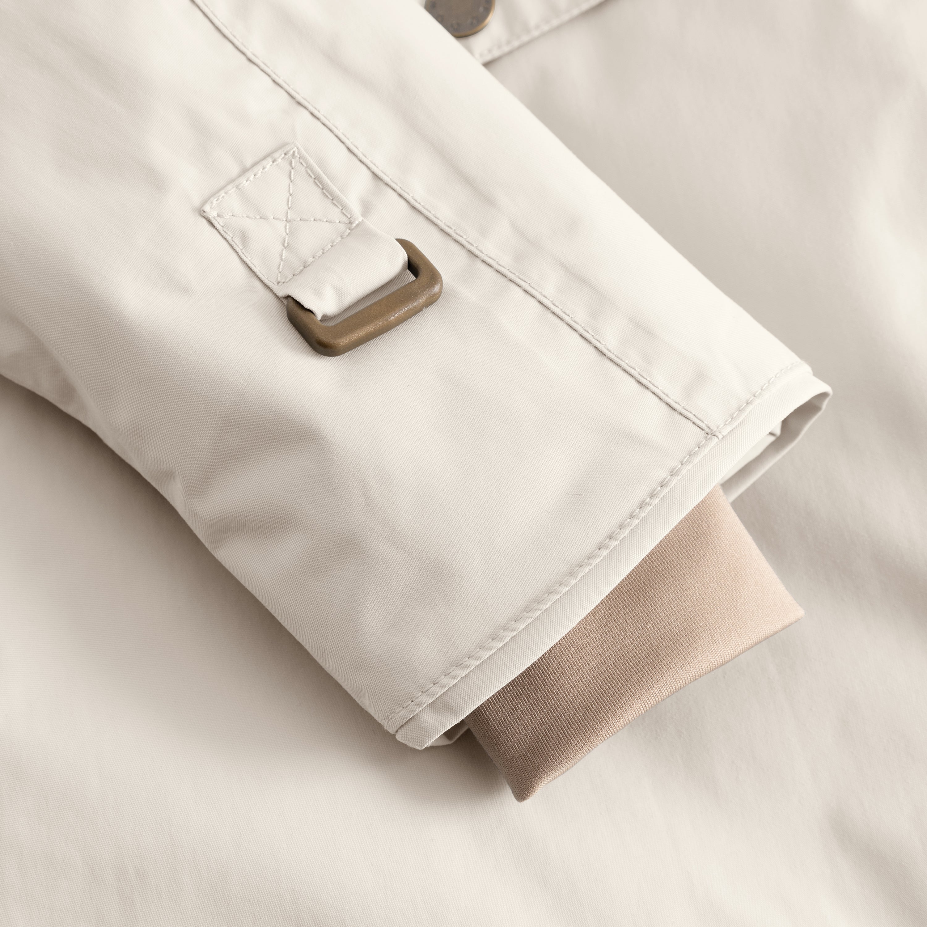 Nærbillede af ærmet på en MATWANG fleeceforet vinterjakke i lys cremefarve, set ovenfra. Fokus er på den bronzefarvede D-ring med krydssting på yderstoffet, mens den beige snekant med tommelfingerhul stikker frem under ærmeåbningen. Øverst i billedet ses en enkelt bronzefarvet trykknap.