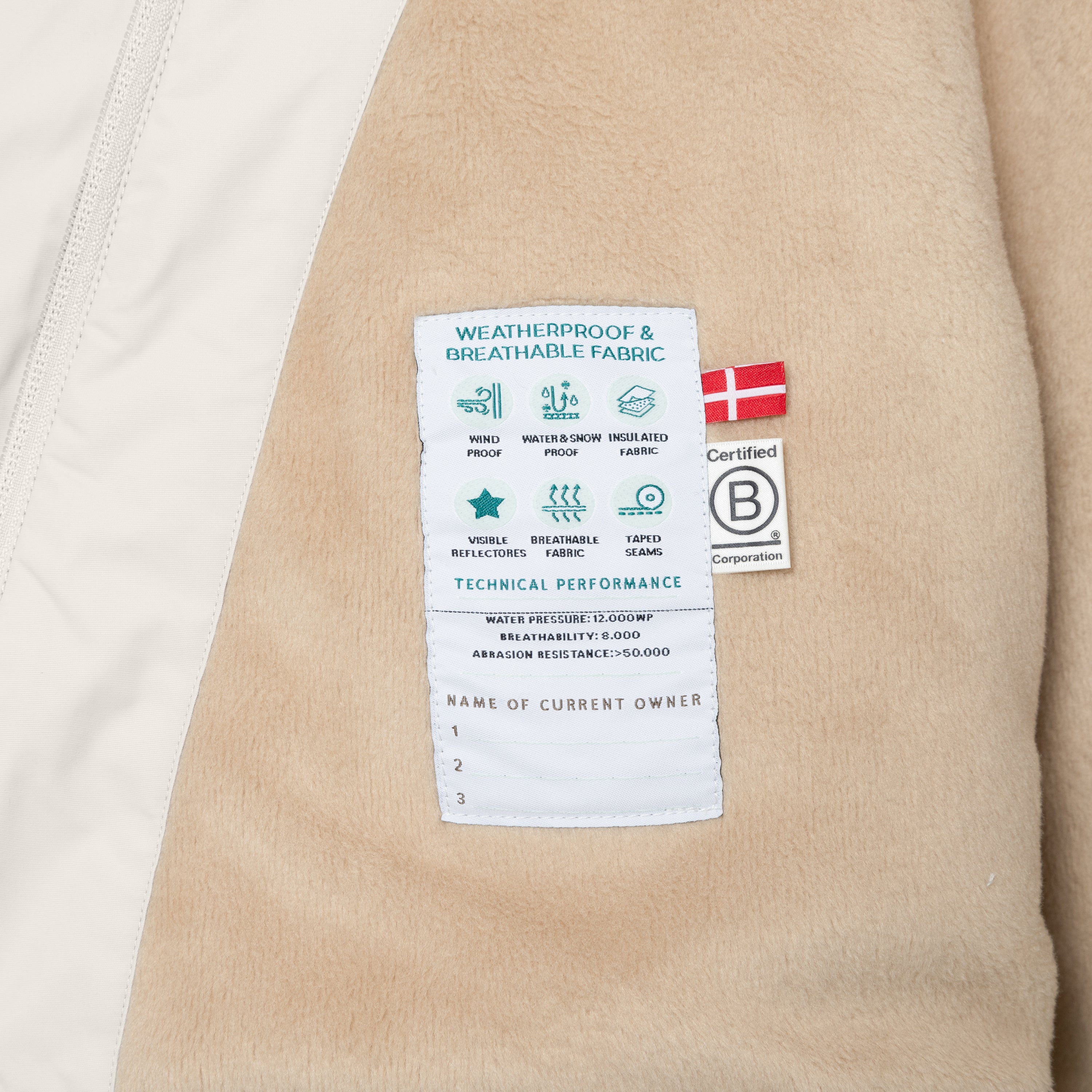 Nærbillede af indersiden af en MATWANG-fleeceforet vinterjakke, hvor et hvidt teknisk mærke med de angivne præstationsværdier for vandtryk, åndbarhed og slidstyrke er syet fast i det bløde, beige fleecefór. Mærket viser ikoner for de vigtigste egenskaber og har plads til at notere op til tre ejere. Det er flankeret af et dansk flag og B Corp-mærket.