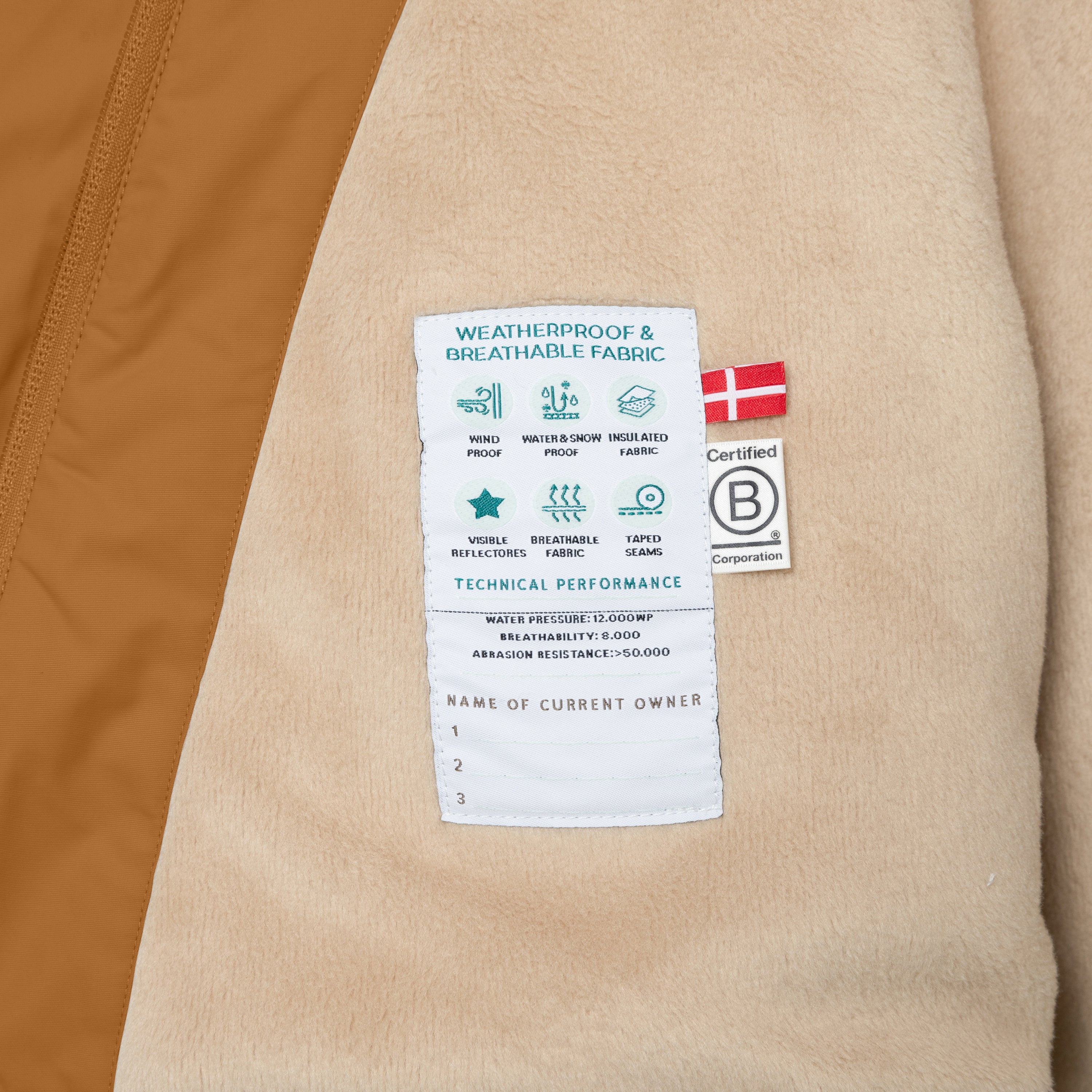 Nærbillede af indersiden af en MATWANG fleeceforet vinterjakke, hvor det bløde beige fleecefór og det tekniske mærke med produktspecifikationer er tydeligt synlige. Her kan du se vandtætheden på 12.000 WP, åndbarheden på 8.000 og slidstyrken på over 50.000, samt et dansk flag og B Corp-mærket, der er syet fast i siden.