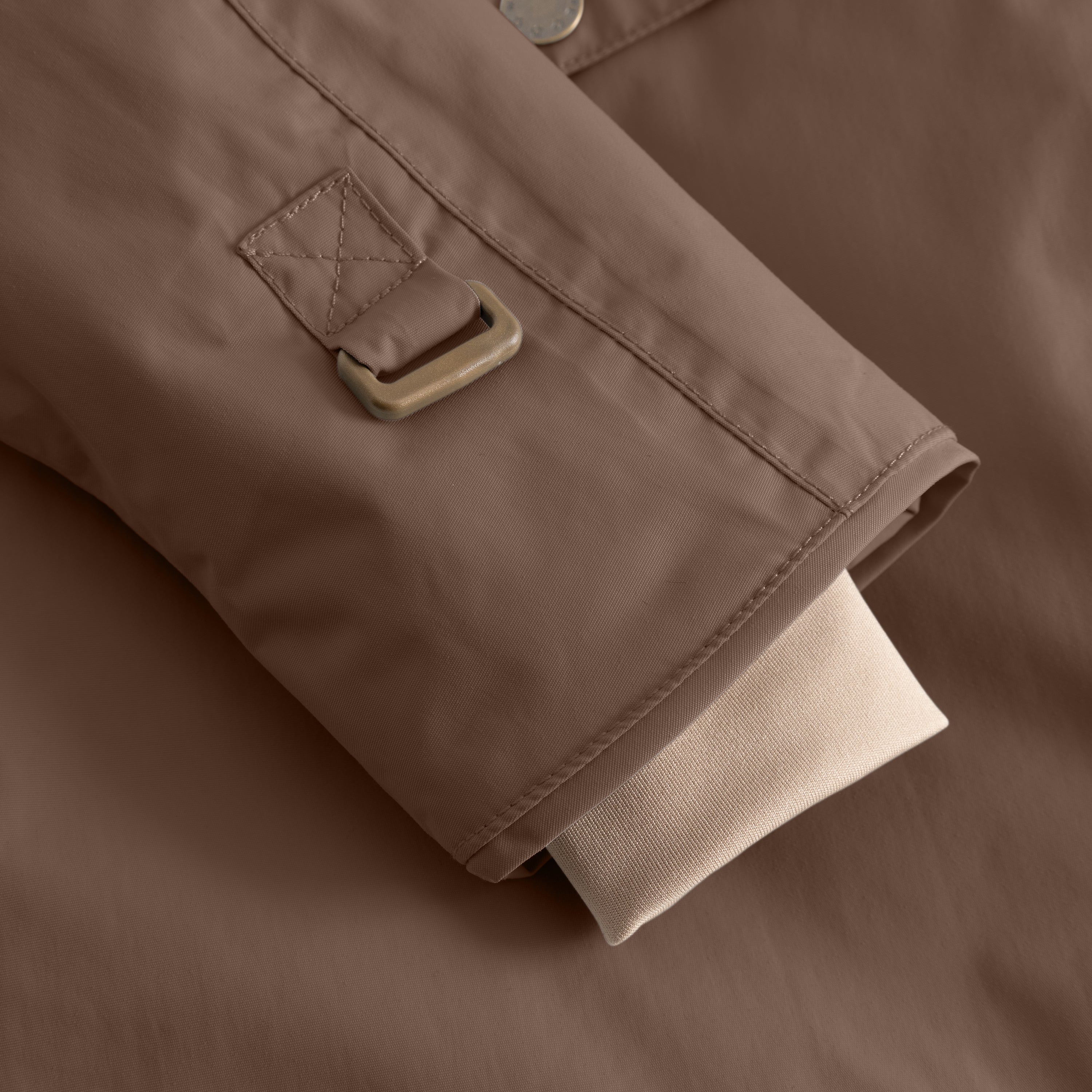 Nærbillede af ærmeafslutningen på en MATWANG fleeceforet vinterjakke i varm gråbrun farve, set fra siden. Den beige snekant med tommelfulds hul stikker frem under ærmeåbningen, mens den bronzefarvede D-ring med krydssting er tydeligt synlig på yderstoffet.