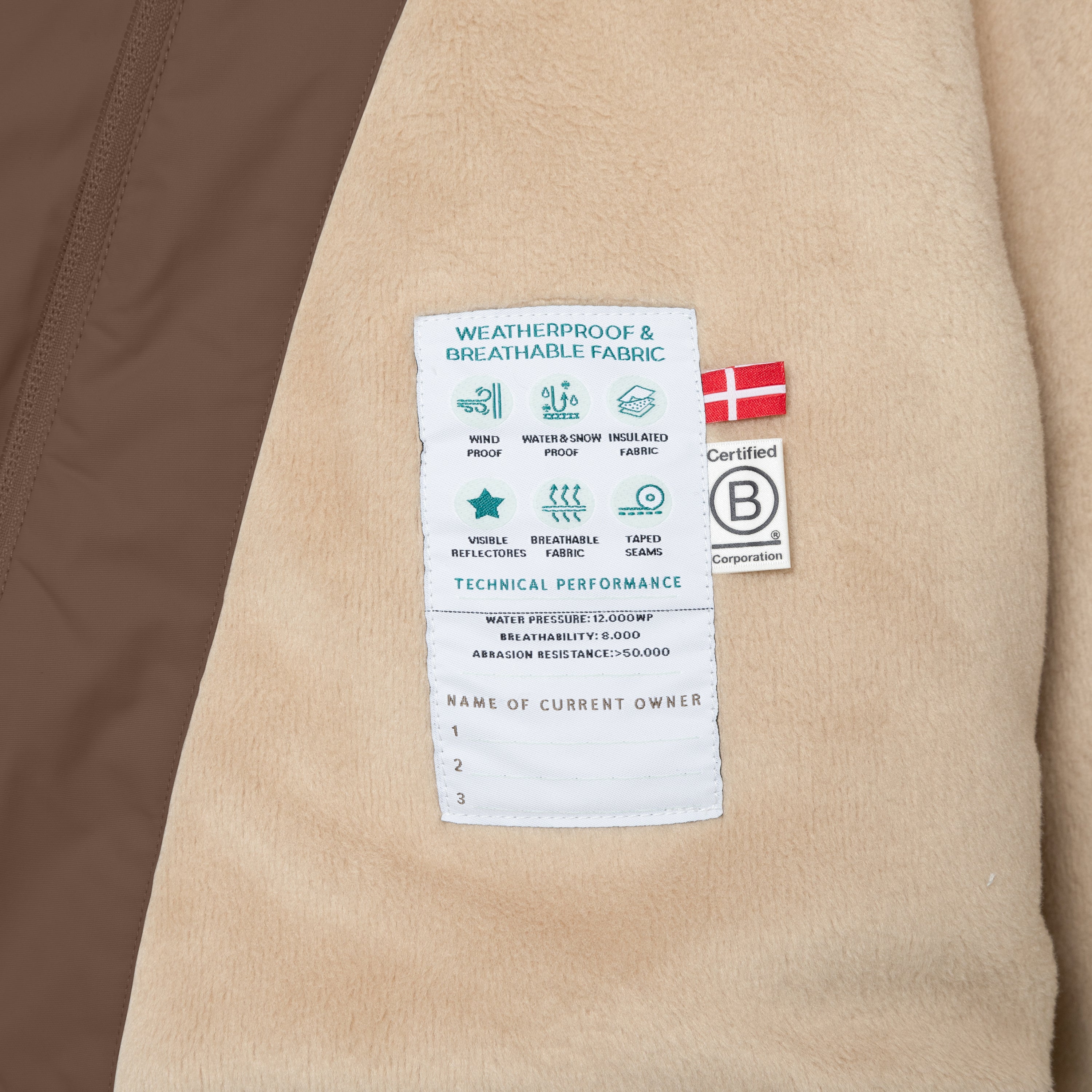 Nærbillede af det bløde, beige fleecefór i en MATWANG fleeceforet vinterjakke. Et hvidt teknisk mærke med ikoner for vindtæthed, vandtæthed, isolering, reflekser, åndbarhed og tapede sømme er syet fast og flankeret af et dansk flag samt B Corp-mærket.