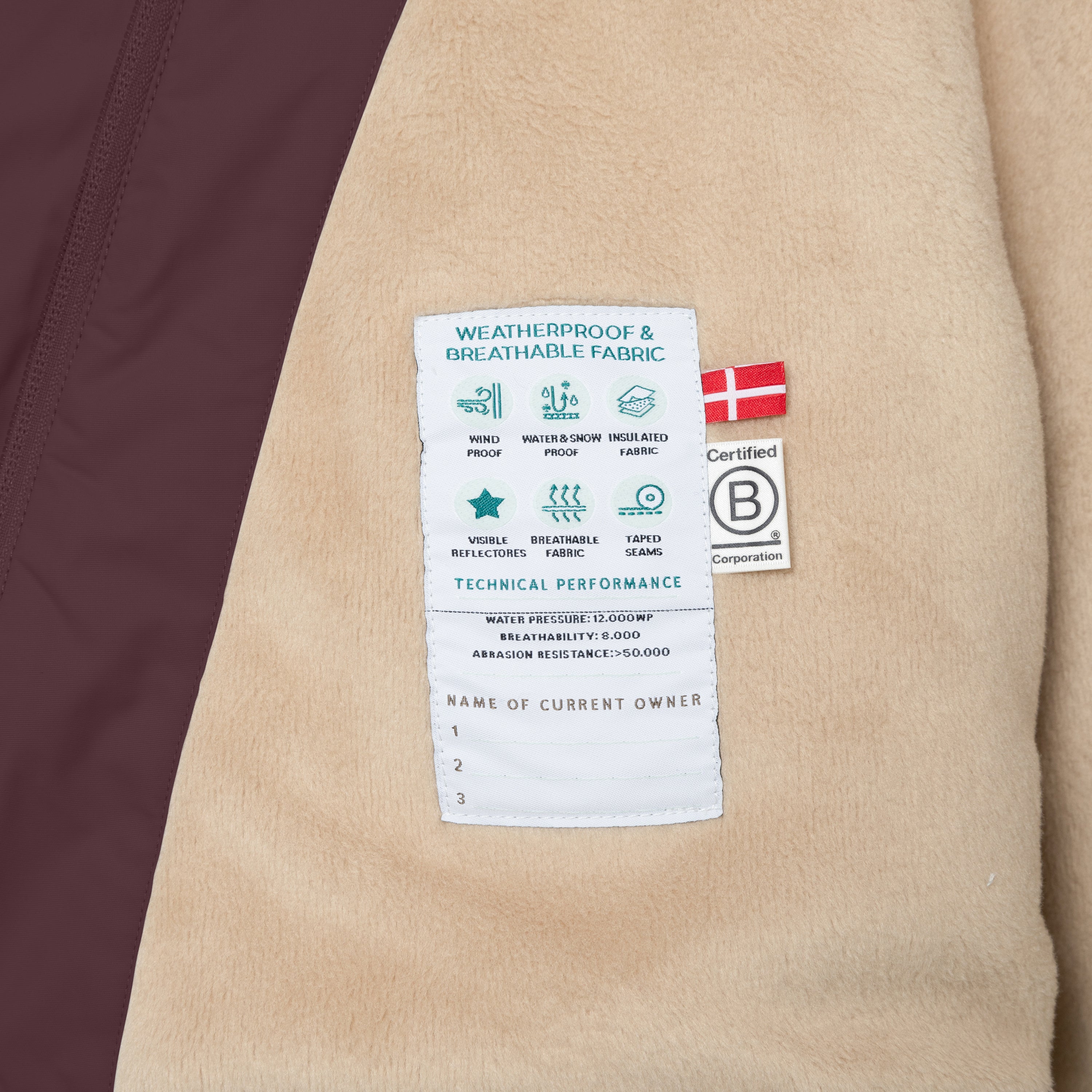 Nærbillede af den tekniske etiket på indersiden af en MATWANG fleeceforet vinterjakke i bordeauxlilla, syet på det bløde, beige fleecefór. Etiketten viser ikoner for vindtæthed, sne- og vandtæthed, isolering, reflekser, åndbarhed og tapede sømme samt de tekniske værdier for vandtryk, åndbarhed og slidstyrke. I hjørnet er et dansk flag og B Corp-mærket synlige.