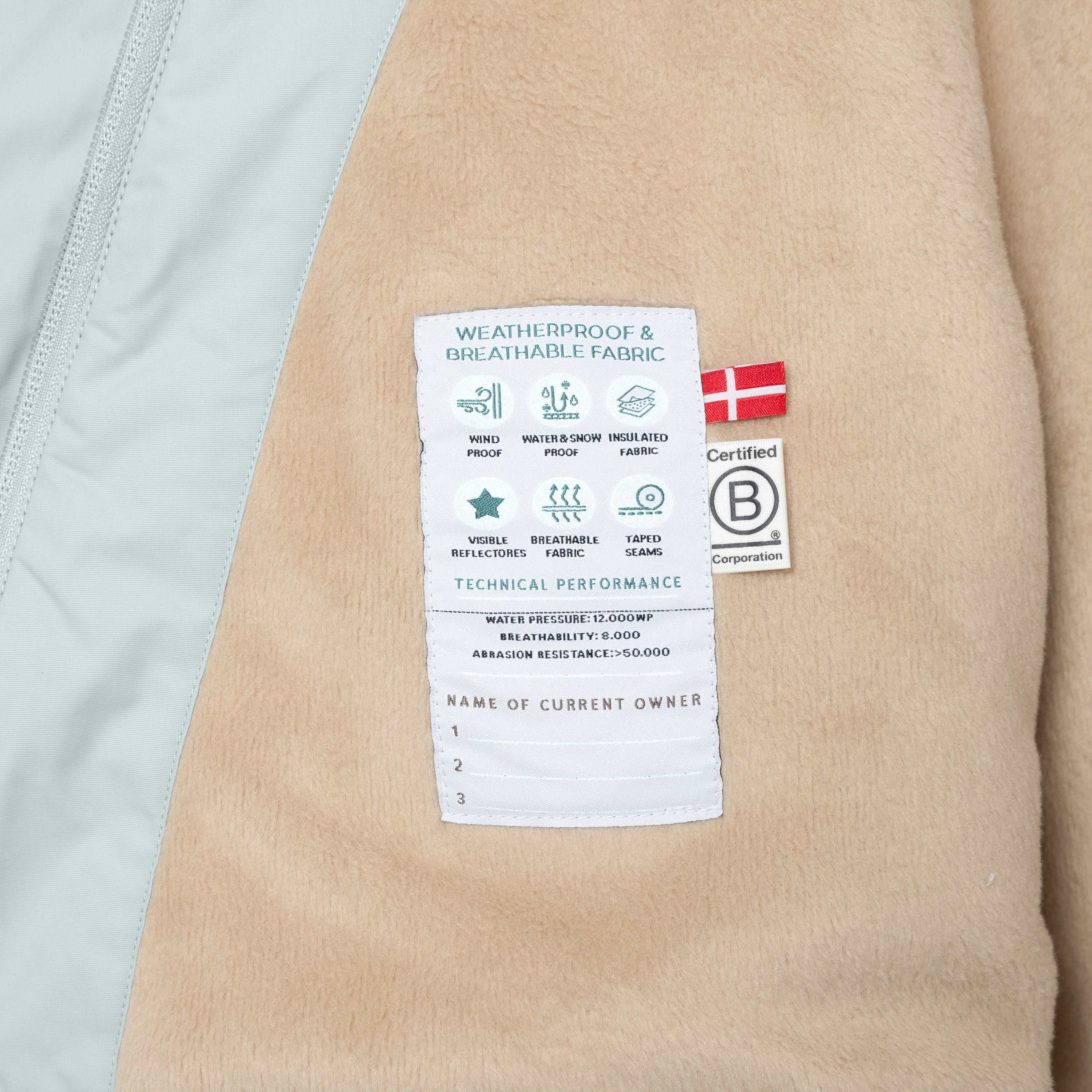 Nærbillede af den tekniske etiket på indersiden af en MATWANG fleeceforet vinterjakke i lys mintgrøn, syet på det bløde beige fleecefór. Etiketten viser ikoner og tekniske værdier for vandtryk, åndbarhed og slidstyrke samt tre linjer til at notere ejerens navn. Derudover ses et dansk flag og B Corp-mærket i siden.