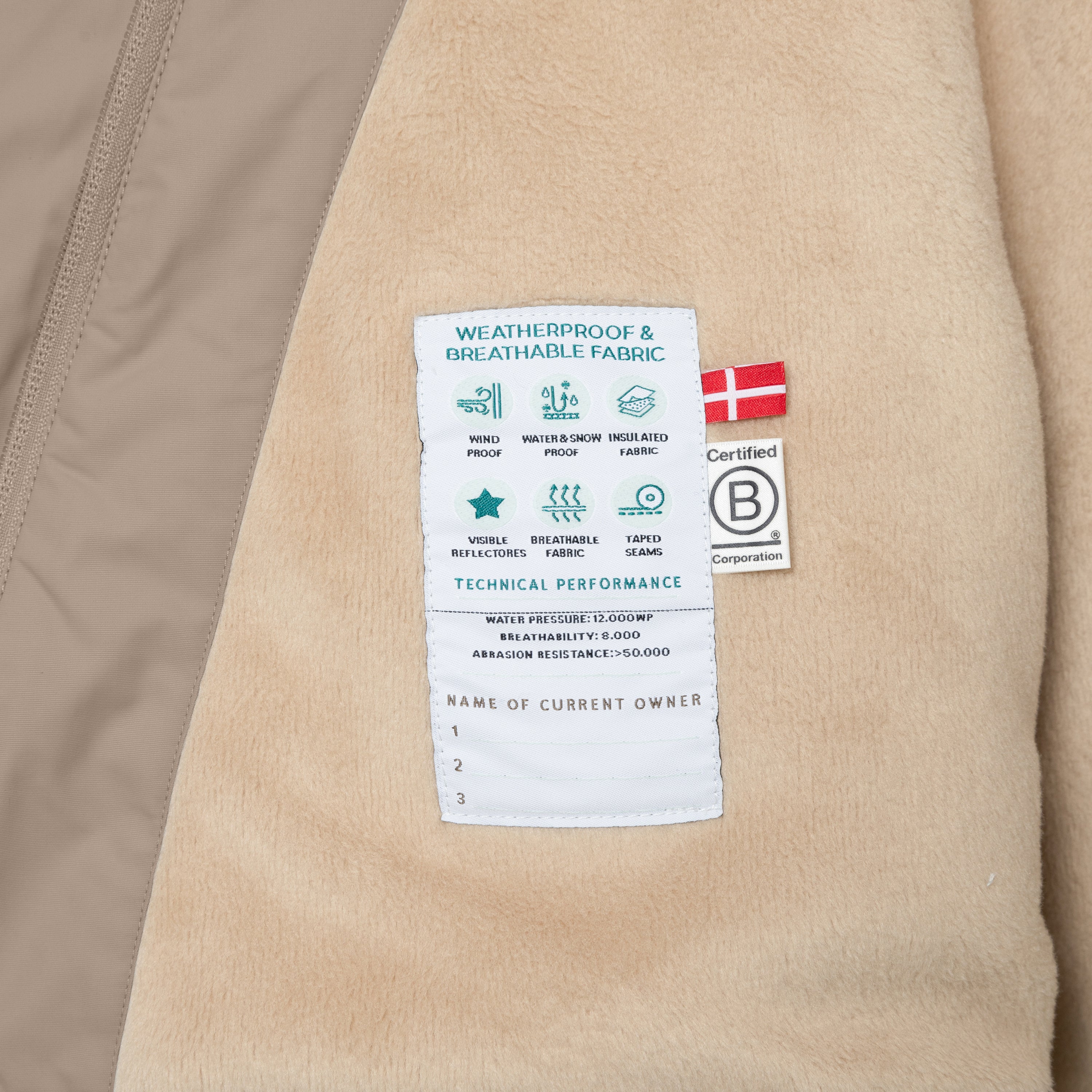 Nærbillede af det bløde fleecefór i en børnevinterjakke med synligt teknisk performancemærke, der viser vandtæthed, åndbarhed og slidstyrke samt et dansk flag og B Corp-certificeringsmærke.
