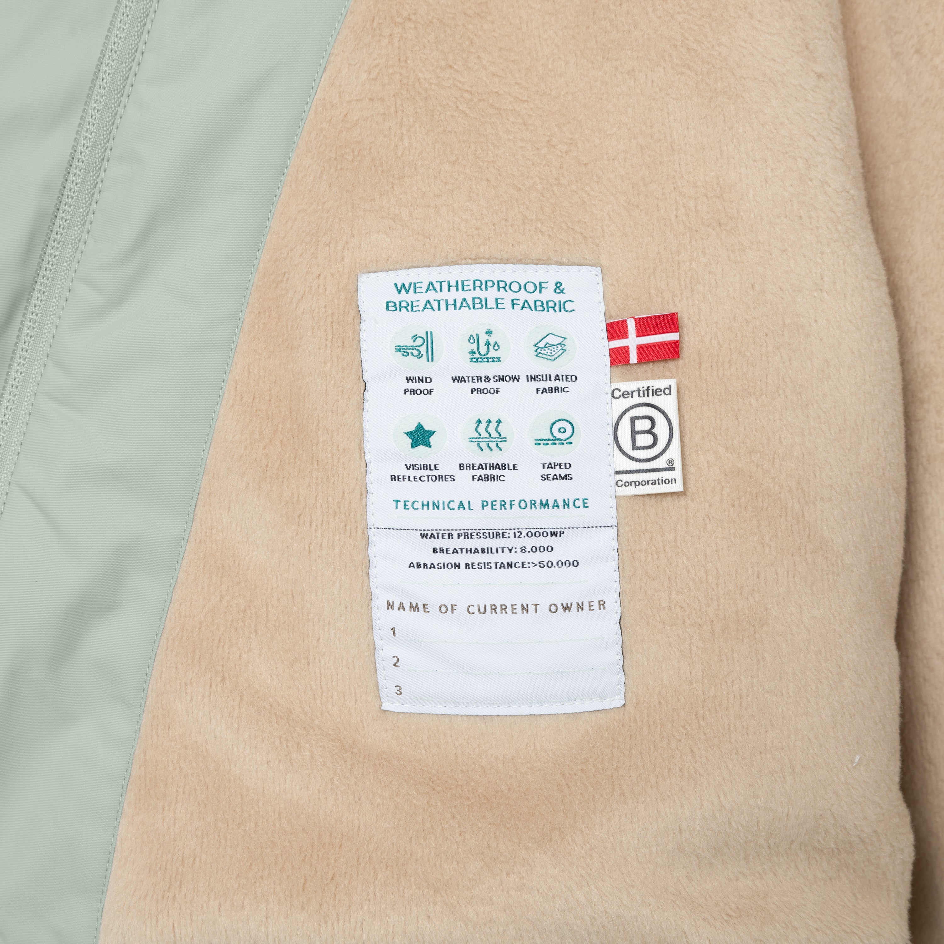 Nærbillede af indersiden af en MATWANG fleeceforet vinterjakke i støvet mintgrøn, hvor en hvid performanceetiket med tekniske specifikationer er syet fast på det beige fleecefór. Etiketten viser piktogrammer for vindtæthed, vandtæthed og snetæthed, isolering, reflekser, åndbarhed og tapede sømme samt et felt til tre ejeres navne. Ved siden af ses et dansk flag-label og B Corp-mærket.