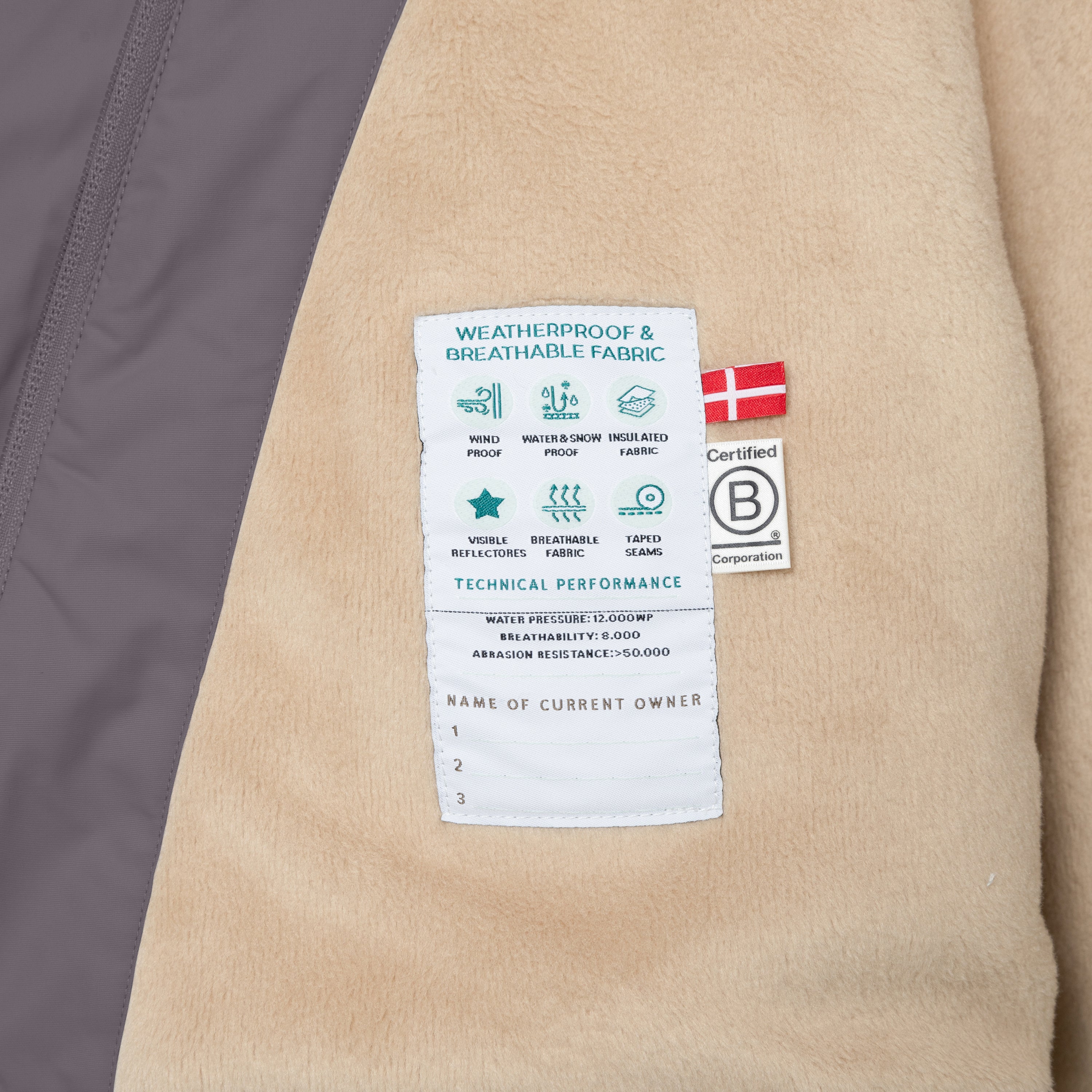 Nærbillede af indersiden af en MATWANG fleeceforet vinterjakke i grålilla, hvor en hvid performanceetiket med tekniske specifikationer og piktogrammer er syet fast på det beige fleecefór. Etiketten viser blandt andet vandtæthed på 12.000 WP, åndbarhed på 8.000 og slidstyrke på over 50.000, og ved siden af ses et dansk flag-label samt B Corp-certificeringsmærket.