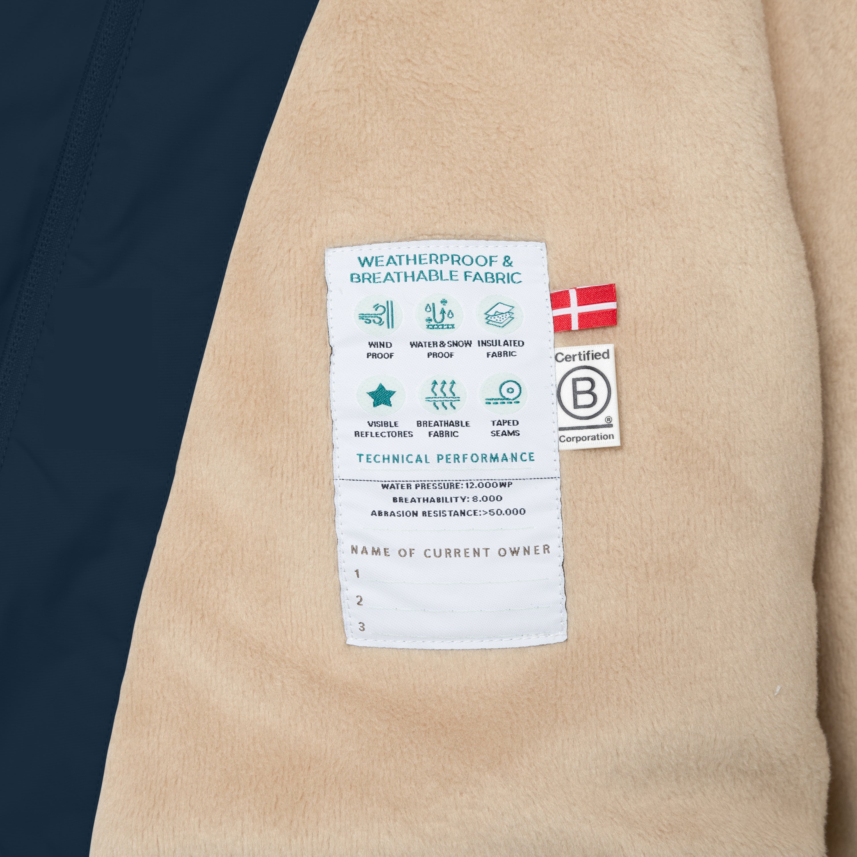 Nærbillede af det bløde fleecefór i en børnevinterjakke med synligt teknisk mærkat, der viser vejrbestandighed, åndbarhed og tapede sømme. Billedet viser også et dansk flag og B Corp-certificeringsmærket.