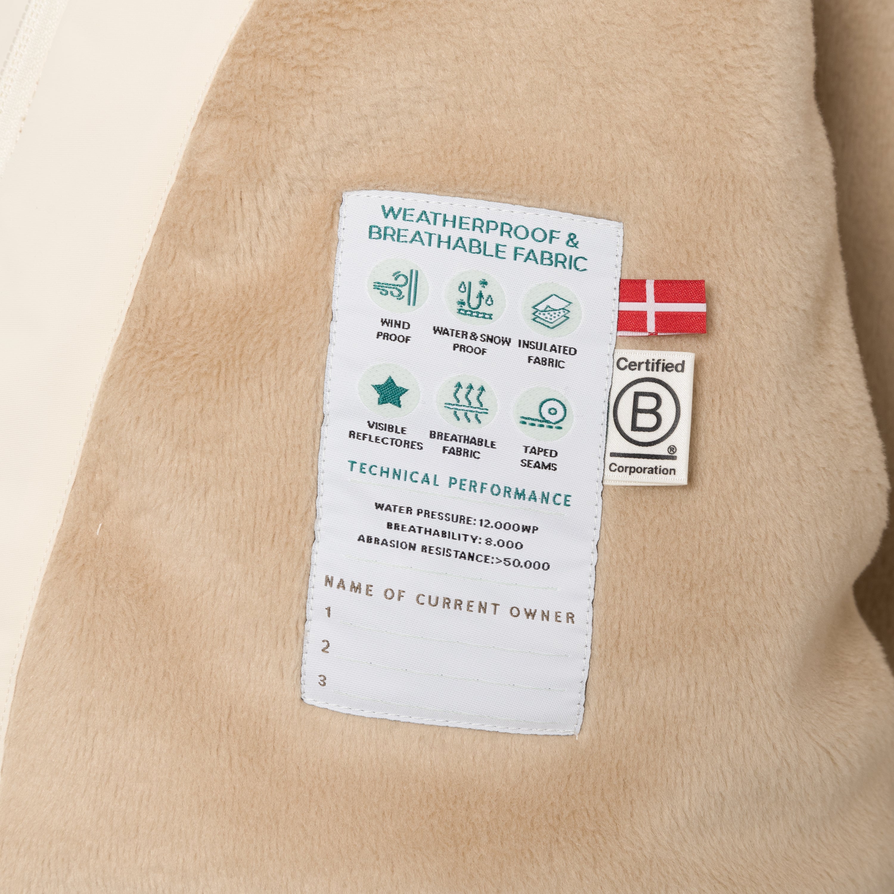 Detaljebillede af det indvendige performancelabel i den cremefarvede MATWANG-fleeceforede vinterjakke med pels, som er syet ind i den bløde, sandfarvede fleeceforing. Labellet viser tekniske specifikationer for vandtryk, åndbarhed og slidstyrke samt ikoner for vindtæthed, vand- og sneafvisning, isolerende stof, reflekser og tapede sømme. Til højre ses et dansk flag og B Corporation-mærket, og nederst er der tre linjer til at notere ejerens navn.