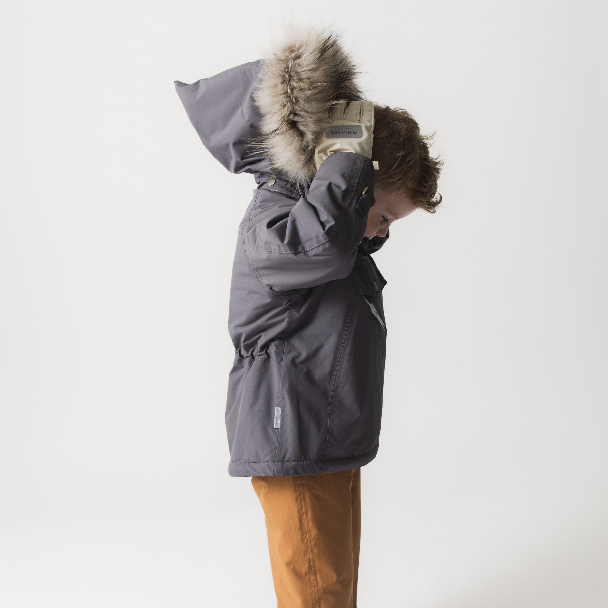 Et barn set fra siden iført en MATWANG fleeceforet vinterjakke med pels i grå, mens det trækker hætten med den store, ægte pelskant op over hovedet. Den beige snekant med tommelfåndshul på ærmet og refleksdetaljen på jakkens forside er synlige.