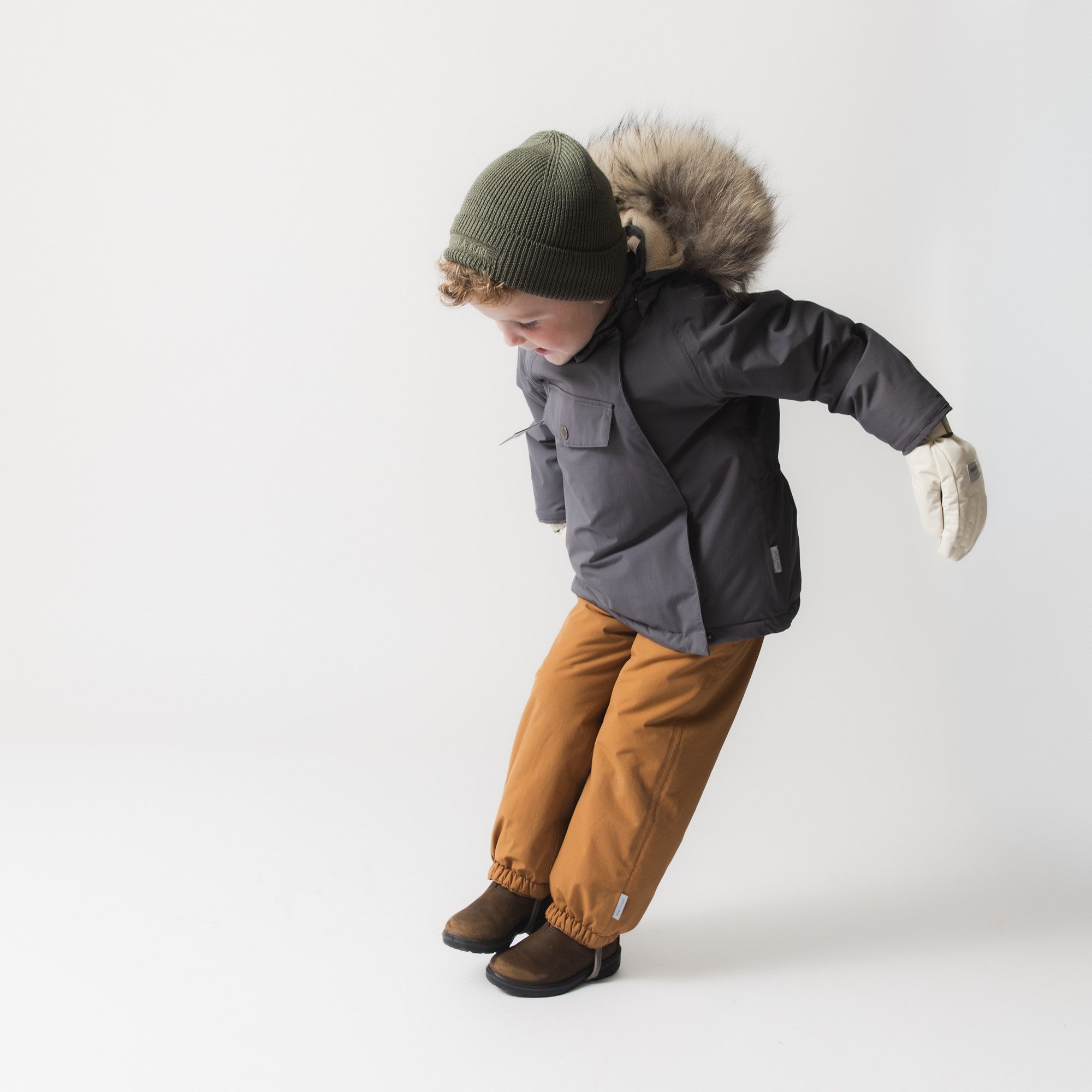 Et barn iført en mørkegrå MATWANG-fleeceforet vinterjakke med pels læner sig frem med armene strakt ud til siderne. Barnet har en grøn strikhue på, beige vanter og brune vinterstøvler. Den store, ægte pelskant på hætten og trykknaplommen fortil er tydeligt synlige.