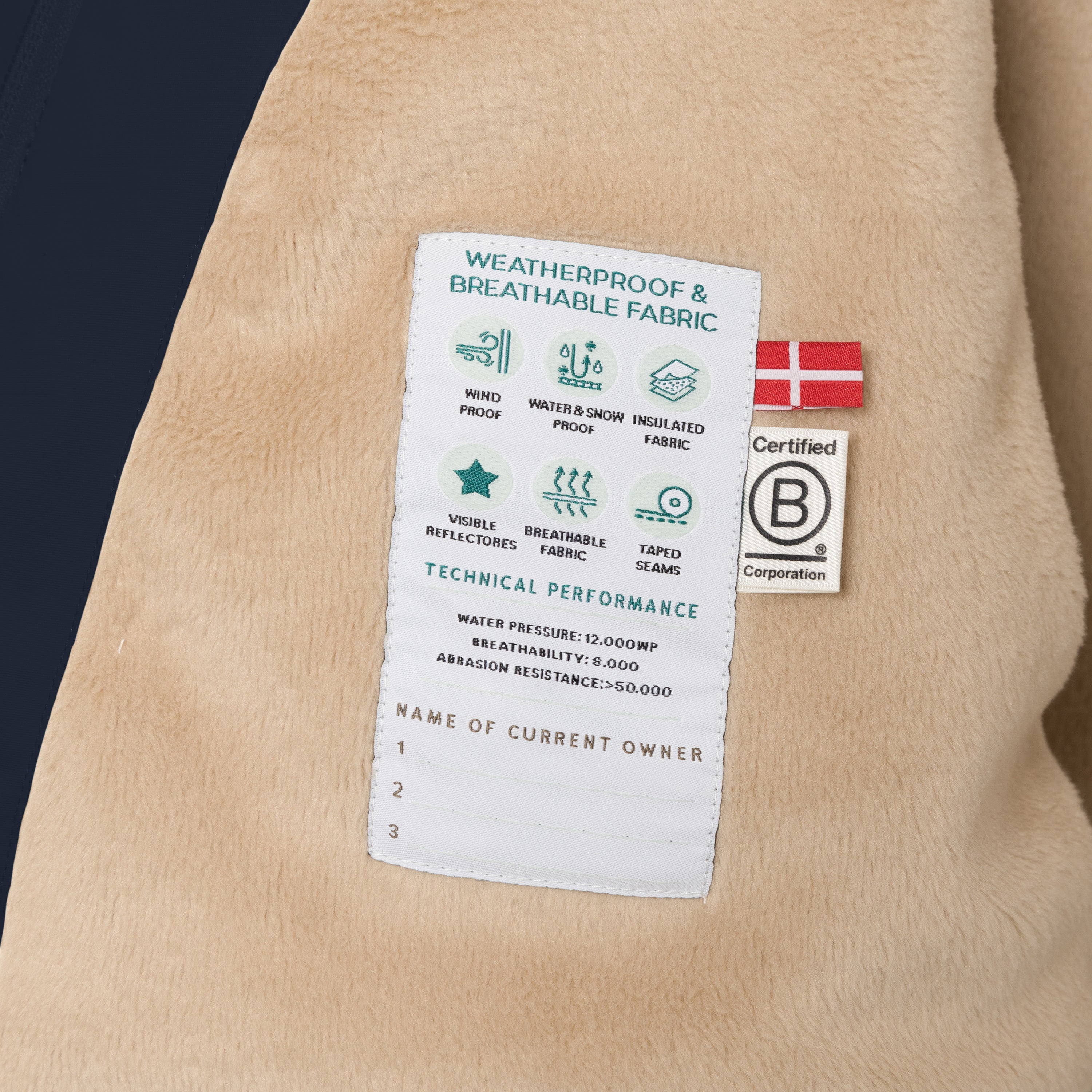 Nærbillede af det indvendige tekniske label i den marineblå MATWANG-vinterjakke med fleecefor og pels. Labellet er syet fast i den bløde, beige fleeceforing og viser ikoner for vindtæthed, vand- og snebestandighed, isolerende stof, reflekser, åndbarhed og tapede sømme samt tekniske værdier for vandtryk, åndbarhed og slidstyrke. Ved siden af ses et dansk flag og B Corporation-mærket.