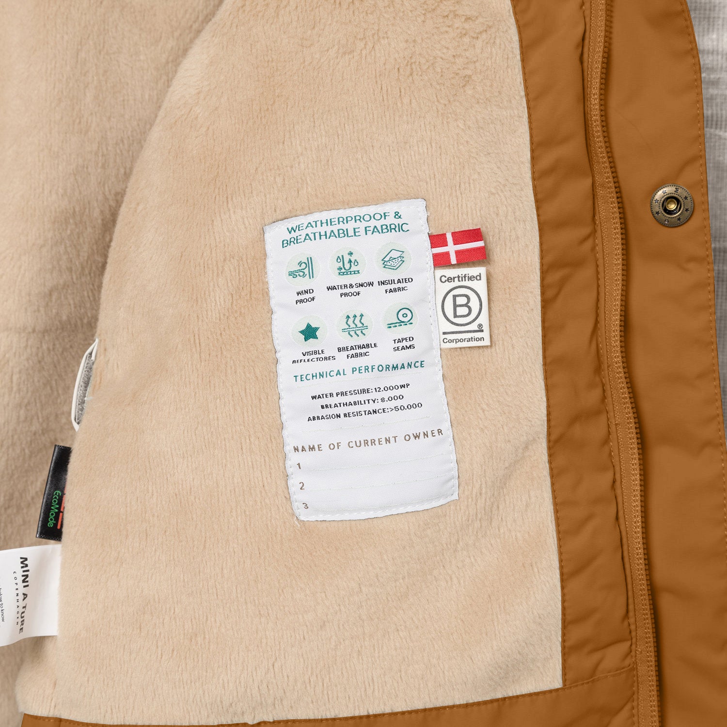 Nærbillede af indersiden af en MATWALLY fleeceforet vinterjakke i karamelfarve, hvor den bløde beige fleeceforing viser en hvid performancelabel med tekniske specifikationer samt B Corp-mærket og et dansk flag. Til venstre ses MINI A TURE- og EcoMade-mærkerne, og langs kanten er det karamelfarvede yderstof med en bronzefarvet trykknap synligt.