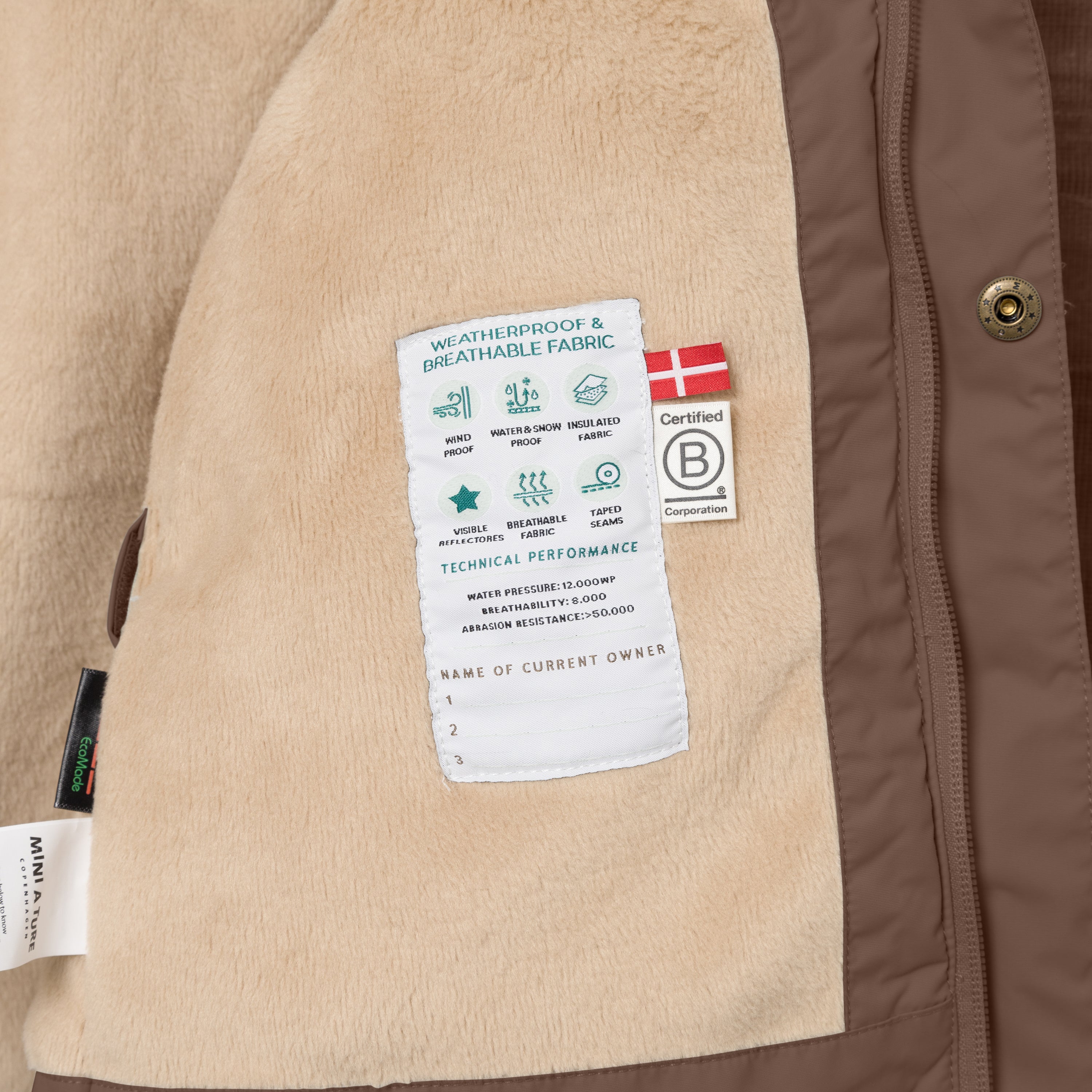 Nærbillede af indersiden af en MATWALLY fleeceforet vinterjakke i varm brun, hvor den bløde beige fleeceforing viser en hvid performancelabel med tekniske specifikationer, et dansk flag og B Corp-mærket samt tre navnelinjer til ejeren. MINI A TURE- og EcoMade-mærkerne ses i venstre side, og en bronzefarvet trykknap er synlig langs den brune lynlåskant til højre.