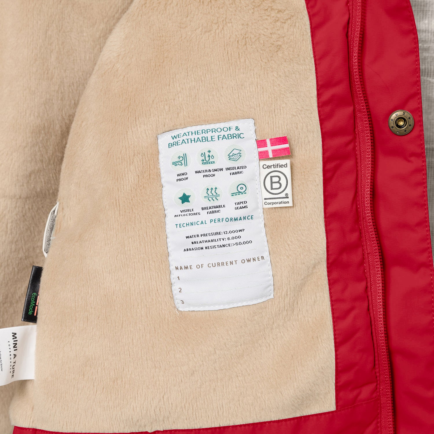 Nærbillede af indersiden af en MATWALLY fleeceforet vinterjakke i klar rød, hvor den beige fleeceforing er åbnet og viser en hvid performancelabel med tekniske specifikationer samt B Corp-mærket. Til venstre ses MINI A TURE-mærket og en EcoMade-label, og langs kanten er det røde yderstof med en bronzefarvet trykknap synligt.