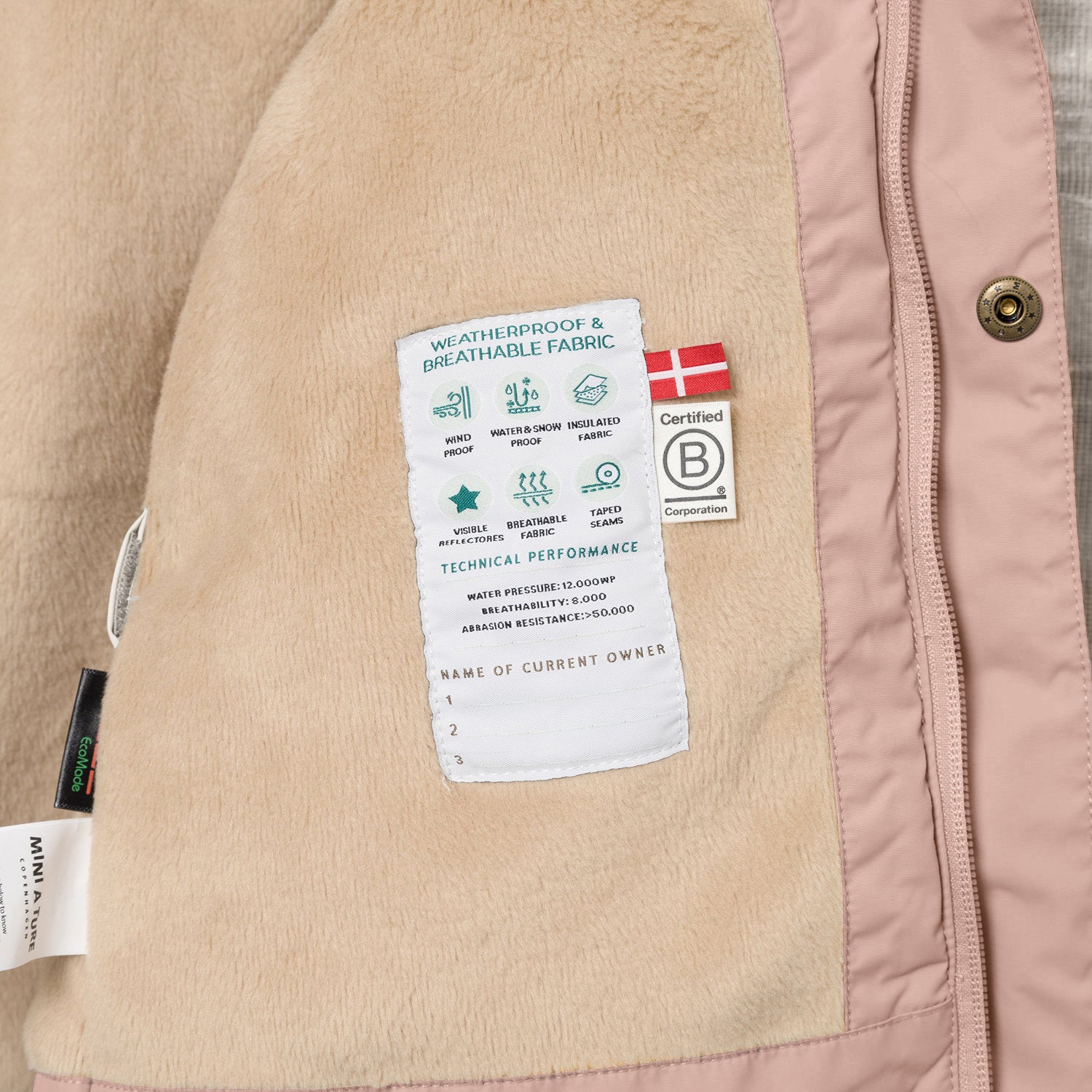Nærbillede af indersiden af en MATWALLY fleeceforet vinterjakke i støvet rosa, hvor den bløde beige fleeceforing viser en hvid performancelabel med tekniske specifikationer, et dansk flag og B Corp-mærket. MINI A TURE- og EcoMade-mærkerne ses i venstre side, og en bronzefarvet trykknap er synlig langs den lyserøde lynlåskant.