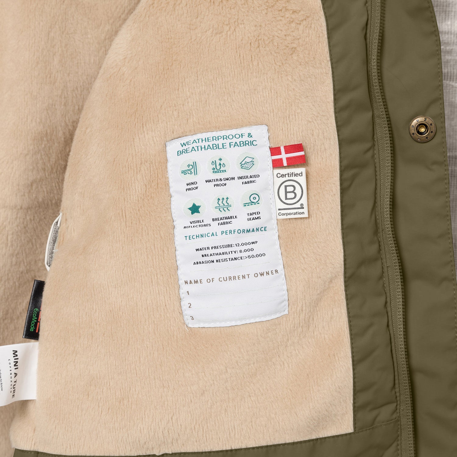 Nærbillede af indersiden af en MATWALLY fleeceforet vinterjakke i olivengrøn, der viser det bløde, beige fleecefor med en indvendig label med tekniske specifikationer, det danske flag og B Corp-certificeringsmærket.