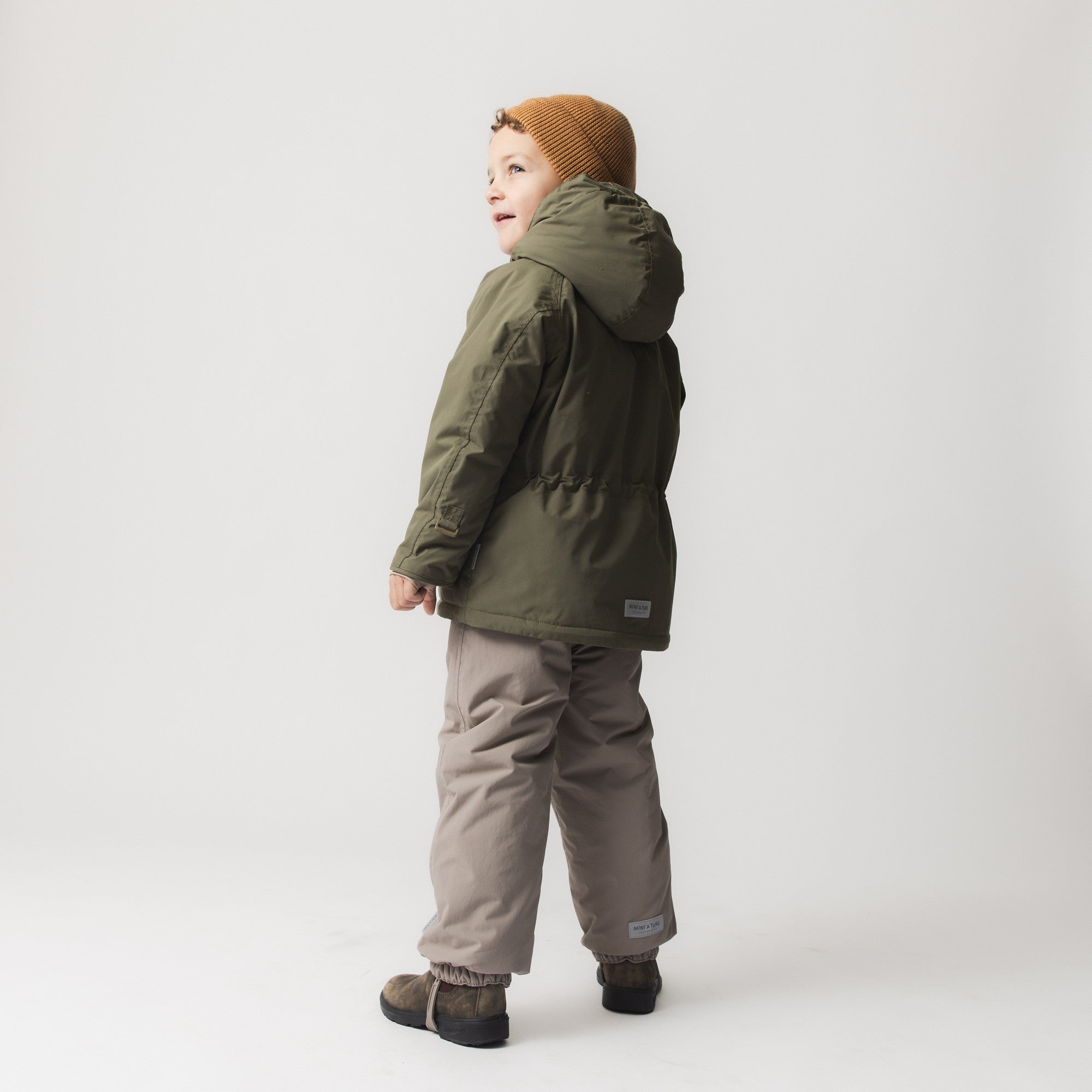 Et lille barn set bagfra i en MATWALLY fleeceforet vinterjakke i mørkegrøn med hætte og MINI A TURE-mærke på ryggen, stylet med lysebeige vinterbukser og en sennepsgul hue.