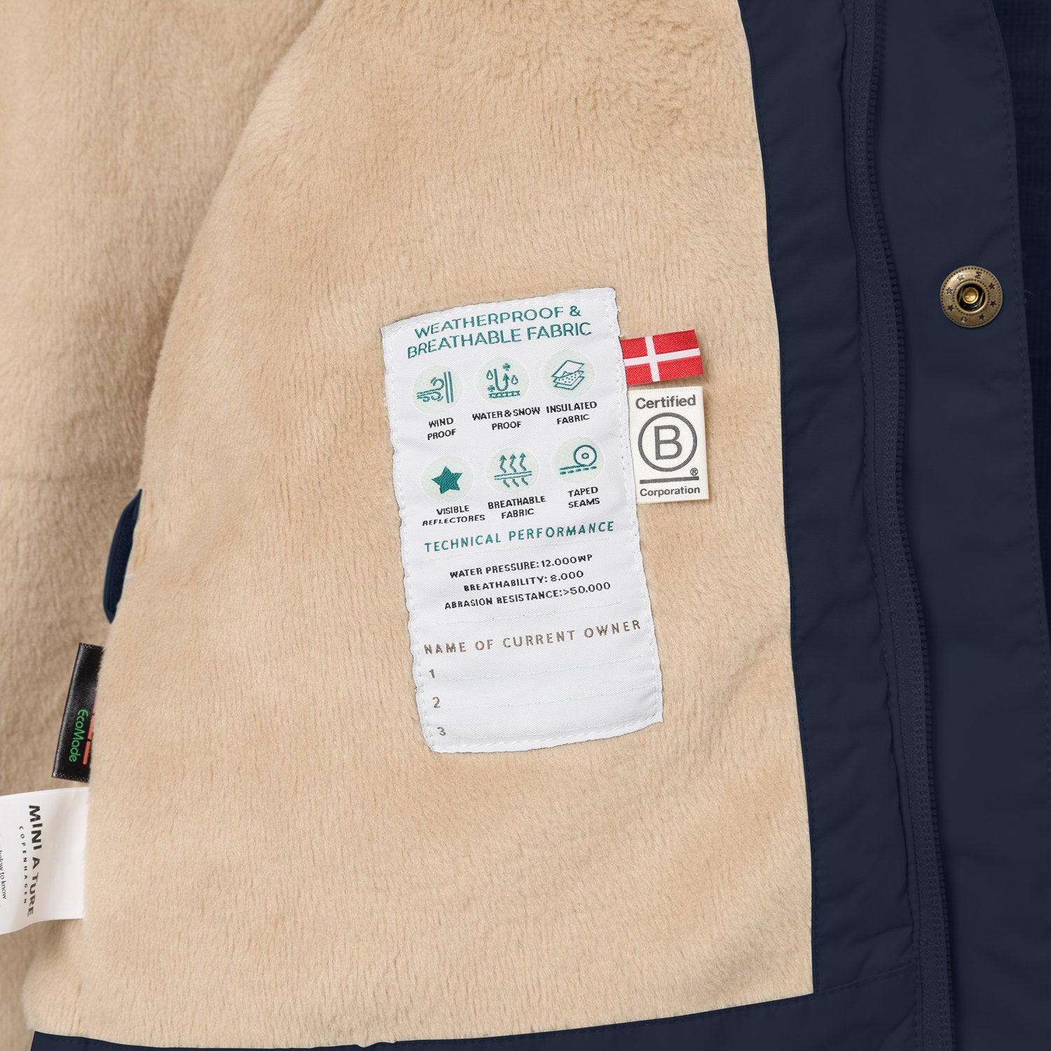 Nærbillede af indersiden af en MATWALLY fleeceforet vinterjakke i mørk marineblå, hvor den bløde, beige fleeceforing ses sammen med en hvid performancelabel. På labelen findes tekniske specifikationer, det danske flag samt B Corp-mærket, EcoMade- og MINI A TURE-labels langs venstre kant.