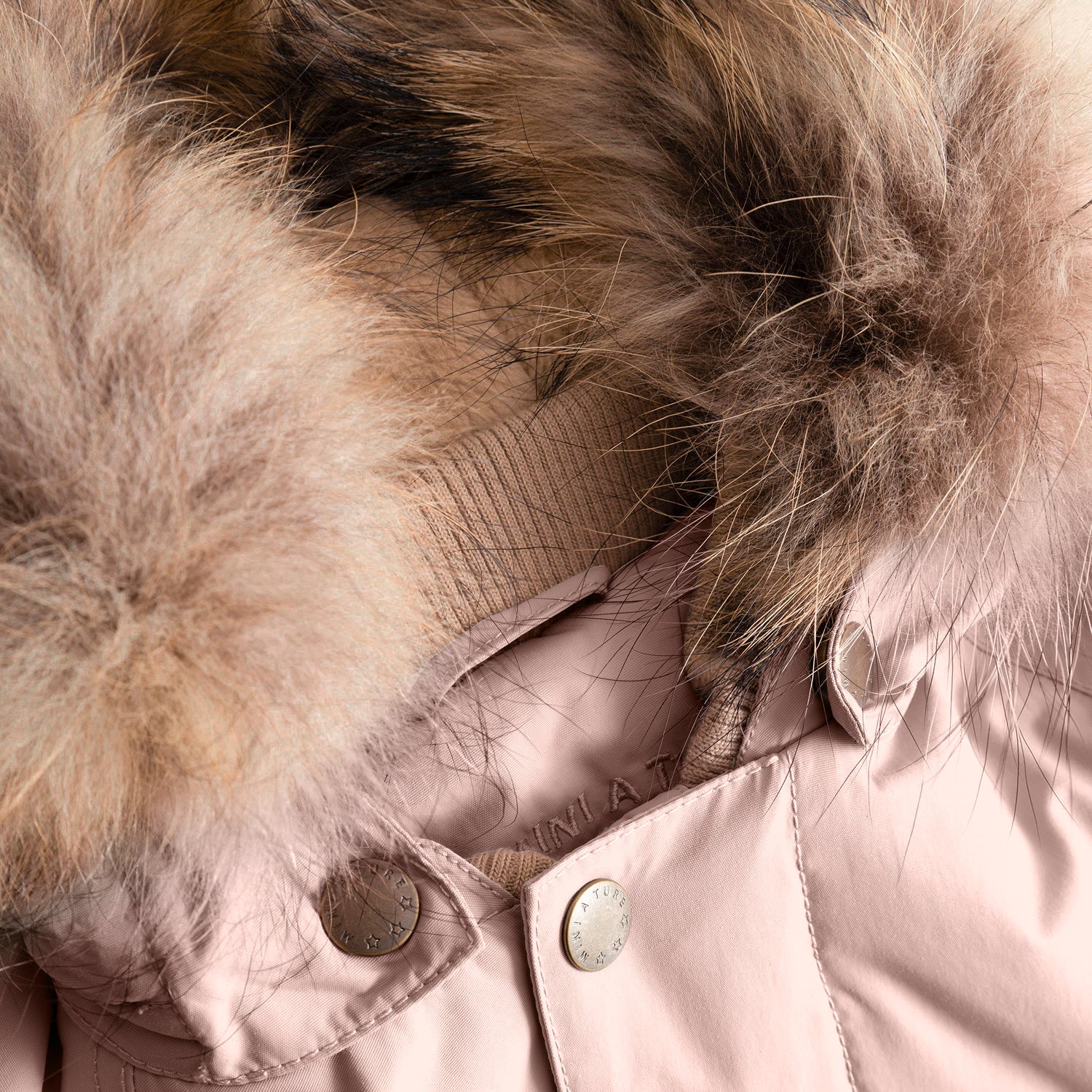 Nærbillede af hætten på en MATWALLY fleeceforet vinterjakke i blød rosa, hvor den voluminøse, naturfarvede ægte pelskant, den beige ribbede halskant og to bronzefarvede MINI A TURE-trykknapper på lukningen ses i tæt detalje.